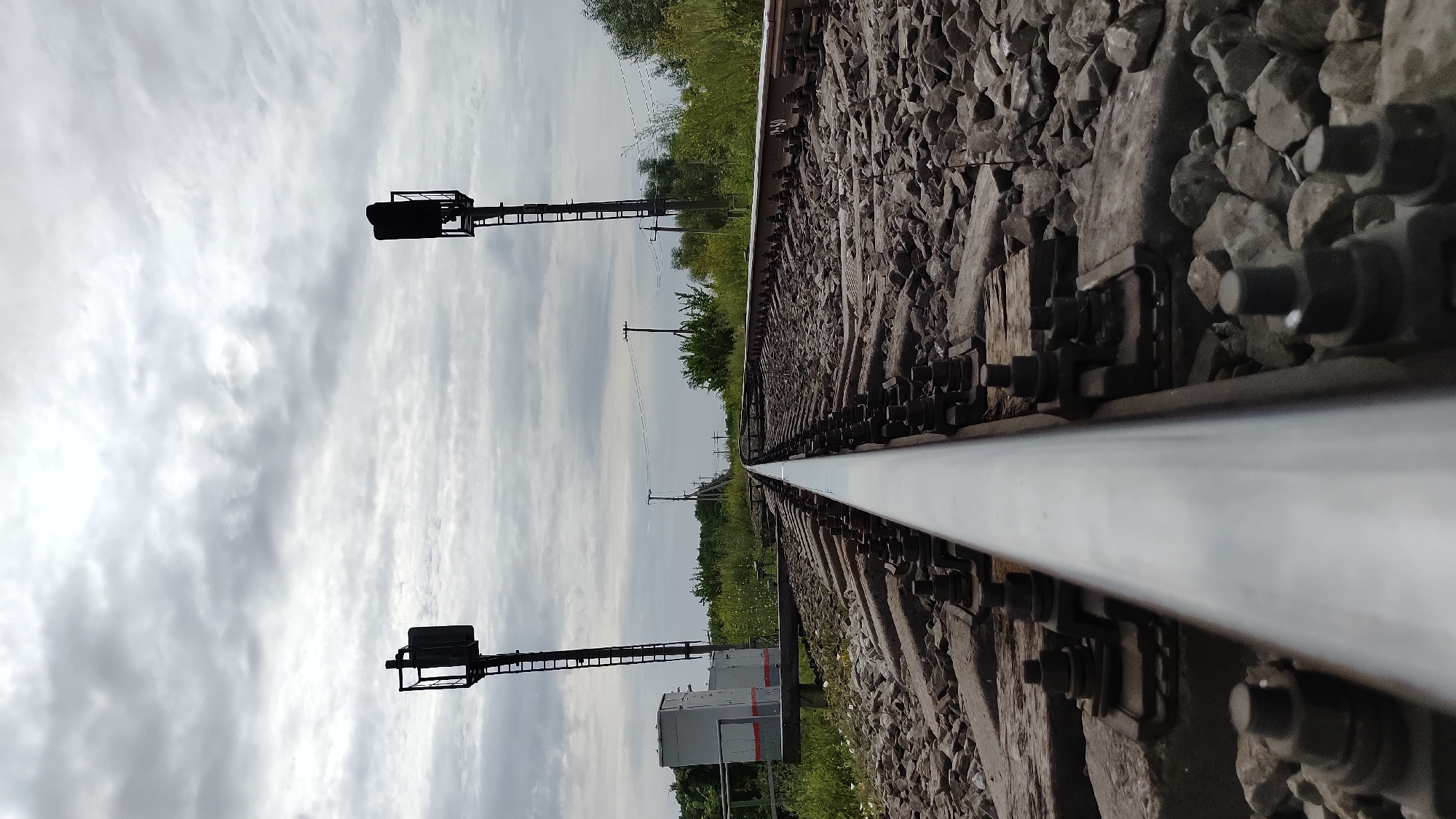 The image is a low-angle perspective shot of a railway track leading into the distance under an overcast sky. Two railway signals with ladders are visible on either side of the tracks. In the foreground, the metallic rail is sharply in focus, with a gravel bed and wooden sleepers beneath it. To the left, a grey electrical box with red stripes is situated next to one of the signals. The background shows a line of trees and utility poles. The scene is set in the city of Mikhaylovka, Russia.