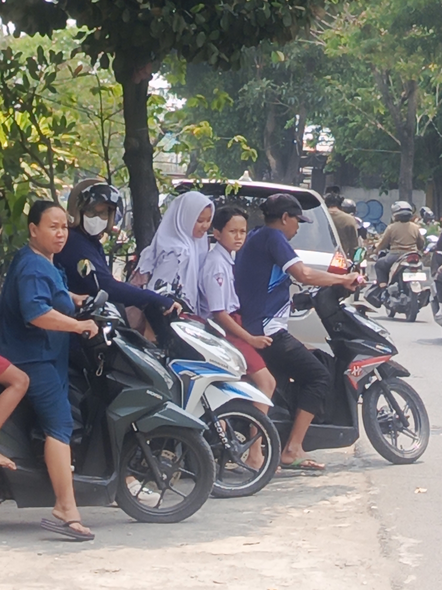 In Surabaya, Indonesia, several individuals are gathered around motorcycles on a paved road with trees and foliage in the background. A woman wearing a blue shirt and dark shorts is seated on a green motorcycle, facing left with her hand on the handlebars. To her right, a person wearing a helmet, mask, and dark long-sleeved shirt is on a black and red motorcycle. Behind them, a young person in a white school uniform with red shorts sits on the same motorcycle, facing forward. Further back, another person wearing a blue shirt and black pants is on a black motorcycle, with a younger individual in a similar white school uniform and red shorts seated behind them. In the distance, other people on motorcycles and a white van are visible. The scene suggests a common mode of transportation in urban Indonesia.