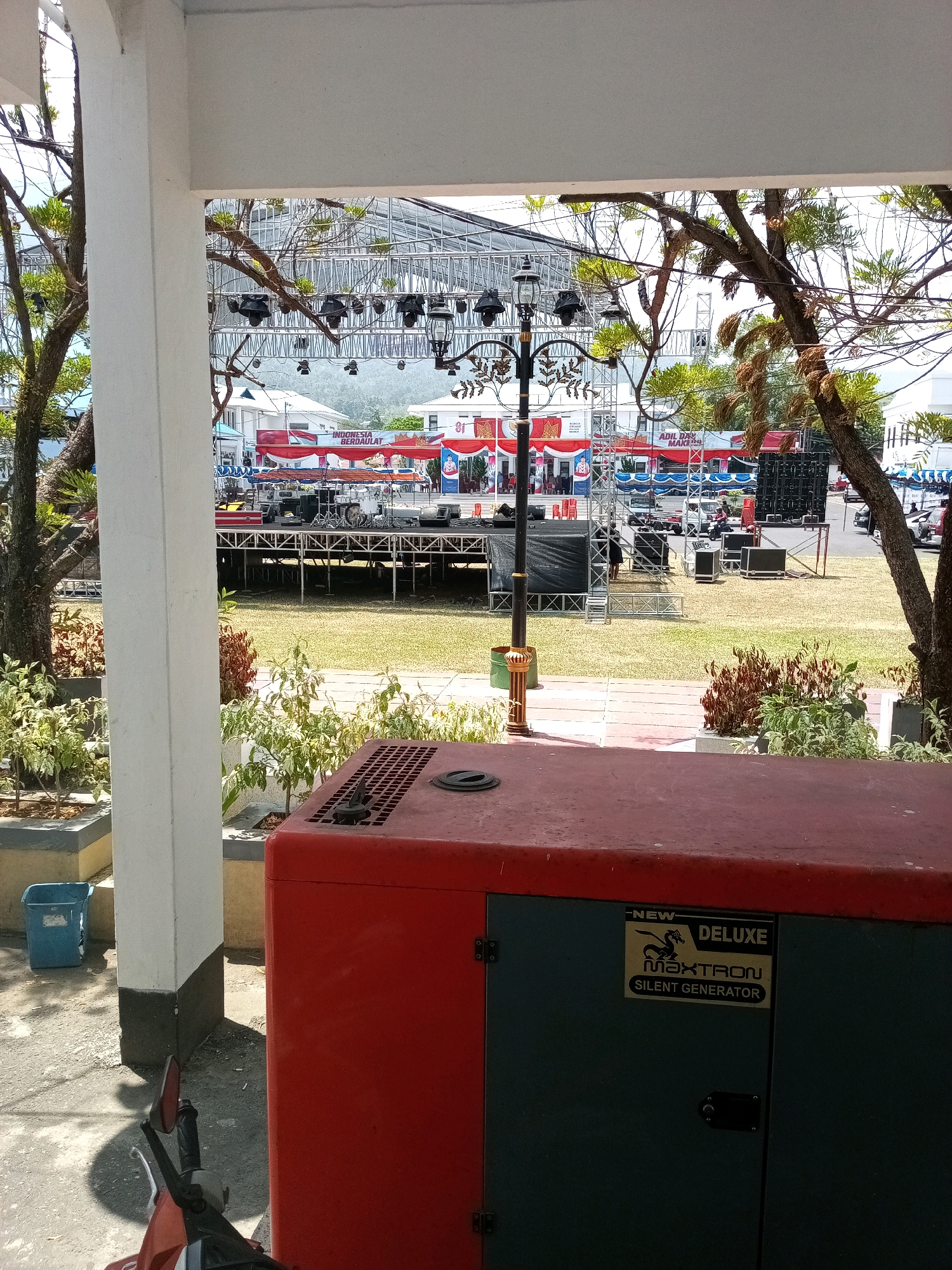The image shows a red and green generator in the foreground, with a black "NEW DELUXE MAXTRON SILENT GENERATOR" label. In the background, there is an outdoor stage setup with lights, speakers, and banners. One banner reads "INDONESIA BERDAULAT 81". The scene appears to be an outdoor event or preparation for one. The location is identified as Bitung, Indonesia, based on the provided context. The environment is grassy with trees and buildings visible in the distance. The lighting suggests daytime.