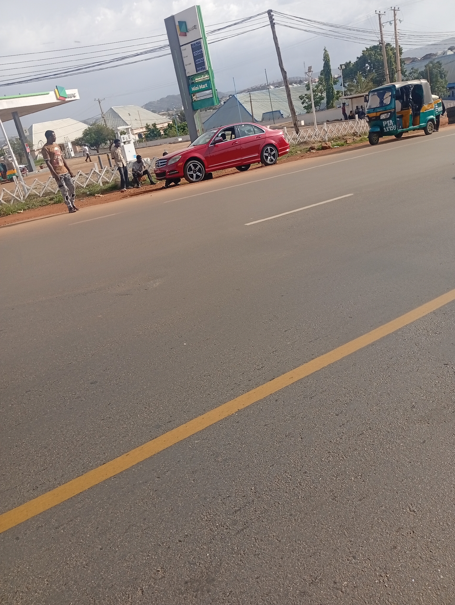 The image depicts a street scene in Rafin Par, Nigeria, under an overcast sky. In the foreground, a paved road is visible, marked with a yellow line on the right side and a white line on the left. A red Mercedes-Benz sedan is parked on the side of the road, facing left. To the left of the car, a man in a brown t-shirt and ripped jeans stands, facing away from the camera. Behind him, near a fuel station, several people are gathered. A blue and green tricycle is parked on the right side of the road. In the background, buildings and trees are present, along with a tall signpost advertising "Mini Mart." Power lines are strung overhead.