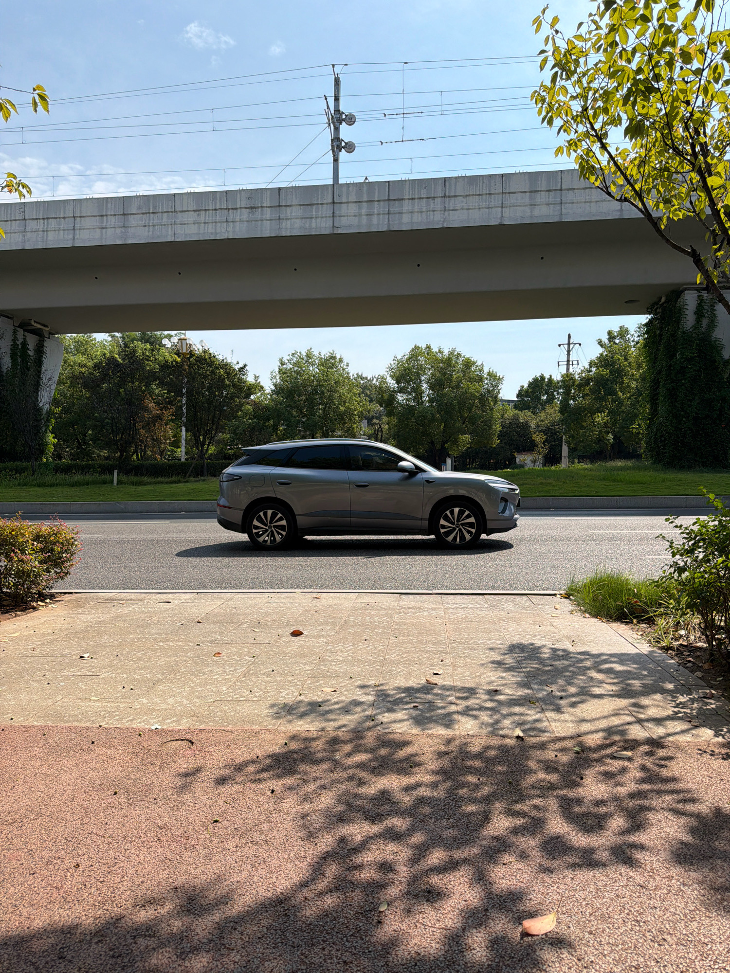 A silver SUV is driving on a road beneath an elevated highway. The sky is clear and blue, with a few clouds. Overhead power lines are visible on the highway structure. Trees and green grass line the side of the road. The foreground shows a sidewalk with patterned paving stones and dappled sunlight from nearby trees. The image appears to have been taken in Jiangjin, China, a district of Chongqing.