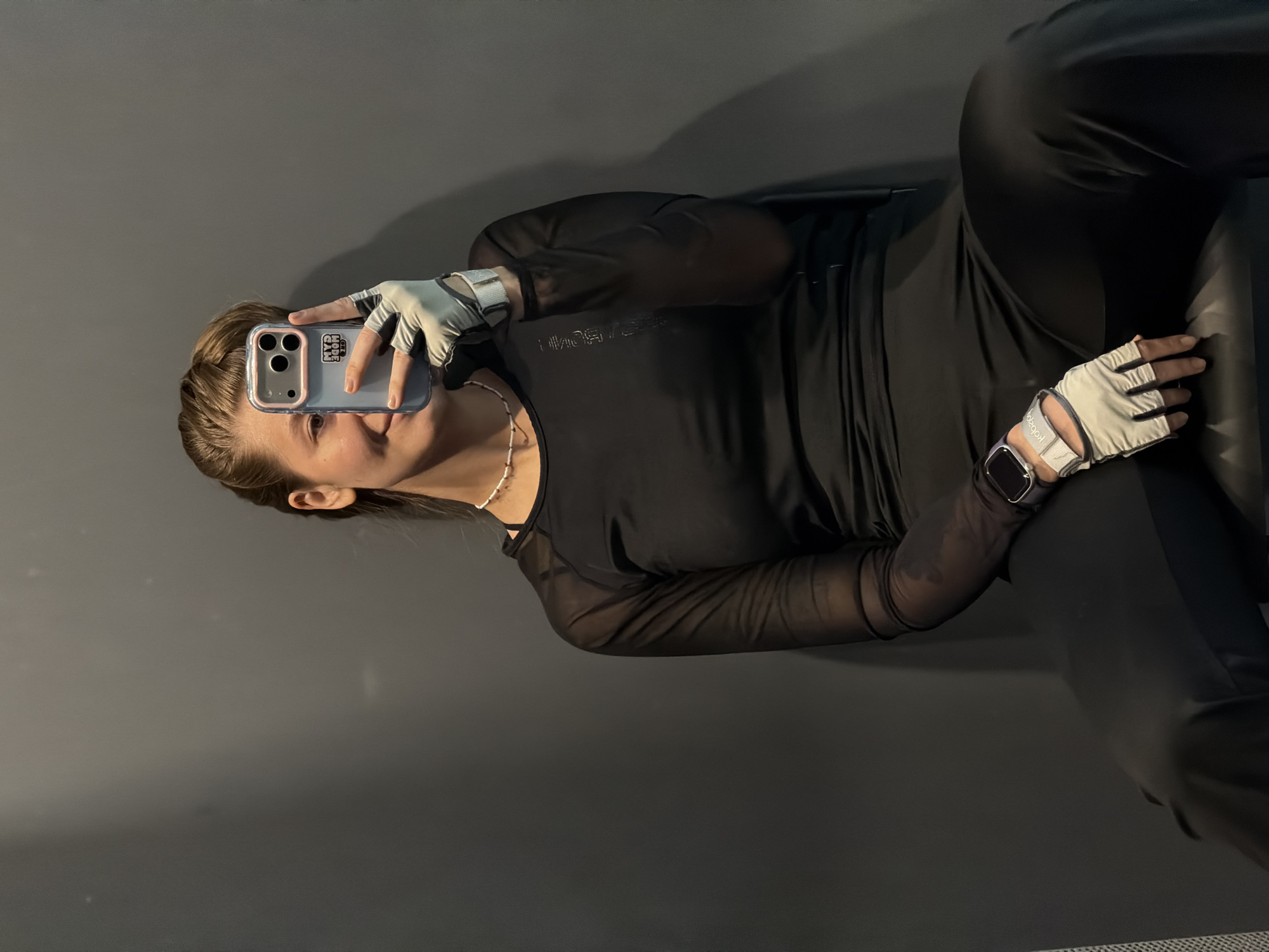 A woman is pictured in an indoor setting, likely in Kazan, Russia. She is positioned in the center of the frame, reclining in what appears to be a chair, and takes a selfie with her smartphone. She has medium-length hair and is wearing fitness gloves. A smartwatch is visible on her left wrist. Her clothing consists of a dark long-sleeved top, paired with dark leggings or pants. The background is a plain grey wall.