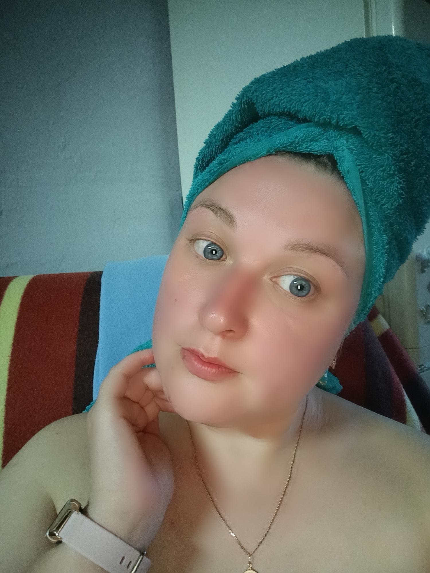 The image is a selfie of a woman with fair skin and blue eyes. She has a green towel wrapped around her head. Her face appears slightly flushed. She is wearing a gold necklace and a watch with a light pink band. Her hand is raised to her cheek. The background includes a multicolored striped fabric and a light blue towel. The location in the city of Chaikovsky, Russia cannot be determined from this image.
