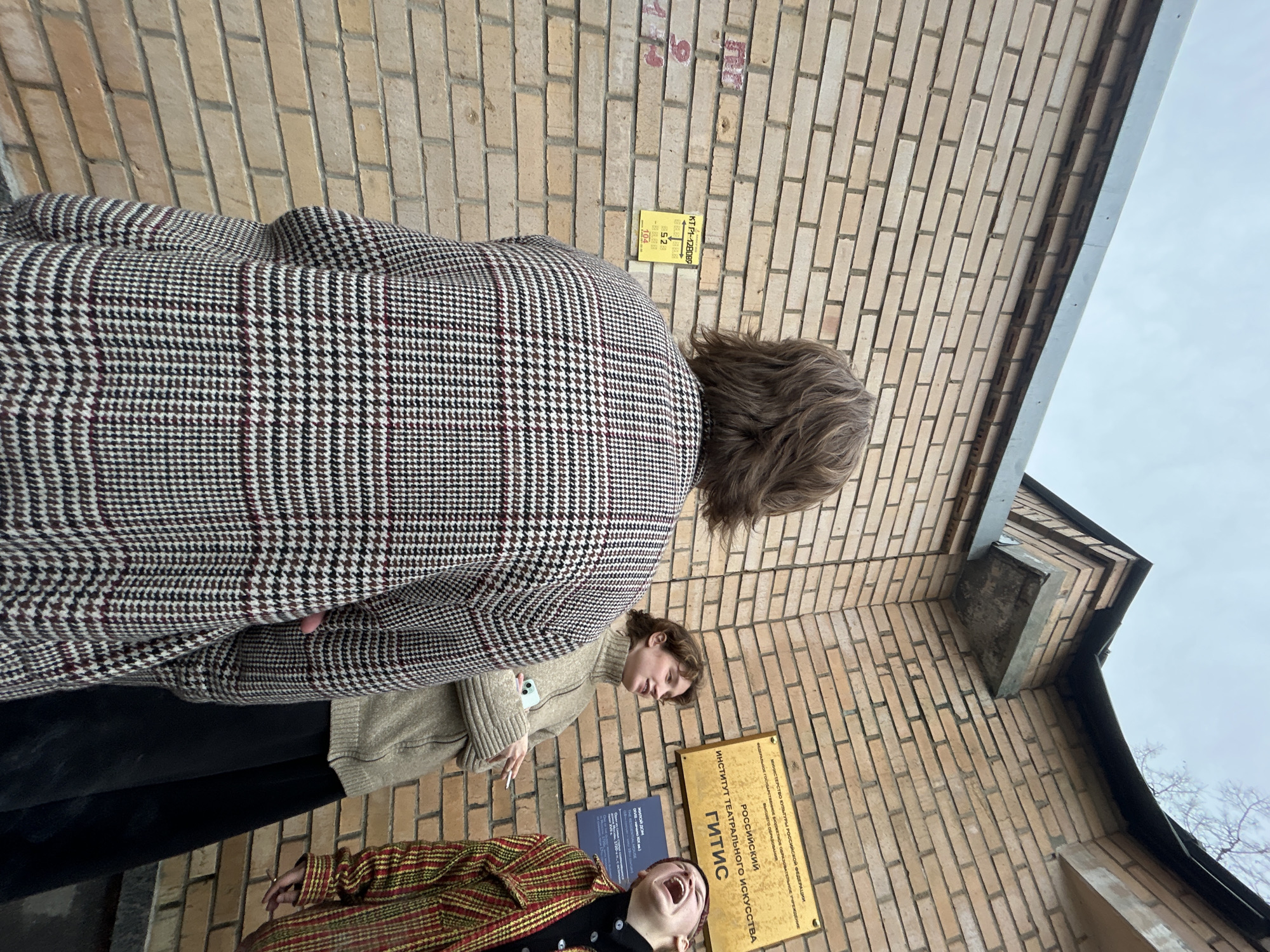Here is a detailed description of the image:
The image captures a scene in front of a brick building, featuring three individuals. One person is primarily in focus, their back turned, wearing a houndstooth patterned coat. Next to them is a person in a beige sweater, and a third individual wearing a red and gold striped jacket is visible at the bottom, appearing to be shouting with their head tilted back.
The setting is characterized by a brick building, with two plaques visible on the wall. One has a small sign that includes text in Russian and numerals. The second larger sign is inscribed with the words "РОССИЙСКИЙ ИНСТИТУТ ТЕАТРАЛЬНОГО ИСКУССТВА ГИТИС" which translates to "RUSSIAN INSTITUTE OF THEATRE ARTS GITIS", further indicating this is likely an educational institution of some kind in an unknown city in Russia. A portion of the sky can be seen in the upper right corner of the frame, suggesting the photo was taken outdoors.