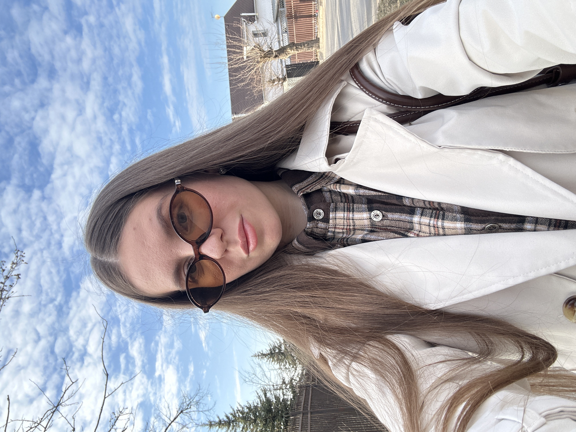 A young woman is depicted in a close-up, likely self-portrait, outdoors in Klin, Russia. She has long, straight, light brown hair parted down the middle and wears oval-shaped, amber-tinted sunglasses. Her lips exhibit a neutral tone. She is dressed in a light beige trench coat over a brown and beige plaid button-down shirt; a dark brown strap, possibly from a shoulder bag, is visible on her right side. The background features a clear blue sky with scattered white clouds. To her left, a dark metal fence separates the foreground from tall coniferous trees and bare deciduous tree branches. To her right, a paved surface abuts a residential area with a two-story white house topped with a dark roof, fronted by a brown wooden fence and a leafless deciduous tree.