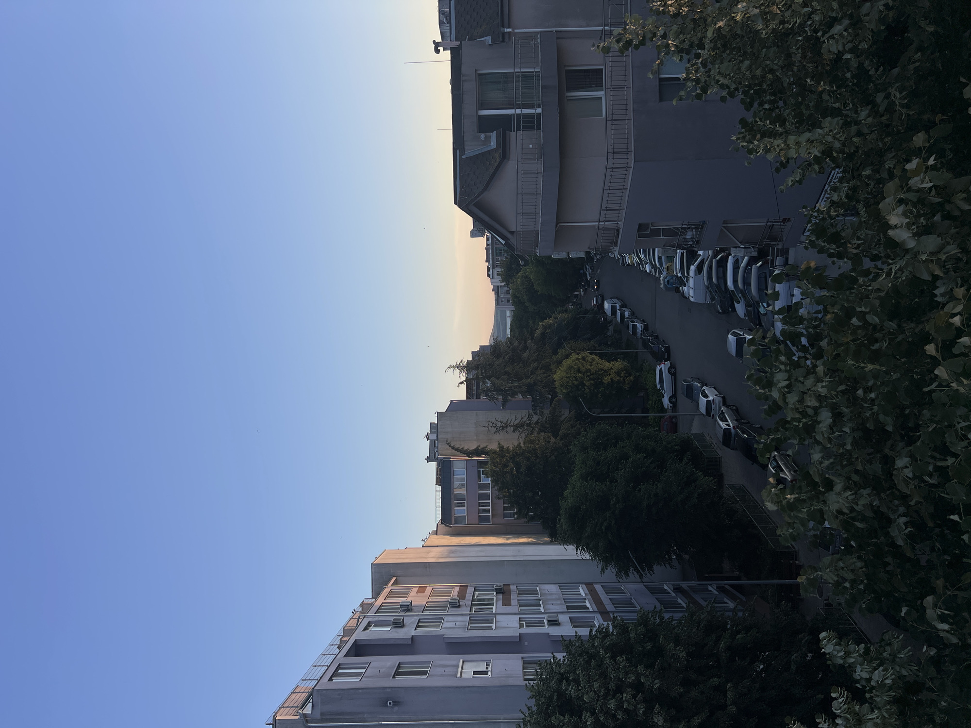 This is a high-angle, full-shot photograph of a street in Burgas, Bulgaria, during the golden hour. The sky is clear and a gradient of blue from light to dark. The sun is setting, casting warm light on the right side of the buildings and street. On the left, a tall, grey residential building with white-framed windows dominates the foreground. To its right, a row of parked cars, mostly sedans and SUVs, line the street. The street is flanked by trees and more residential buildings. A prominent, light-pink building with a gabled roof and balconies stands on the right side of the image. The overall impression is a quiet urban scene at dusk.