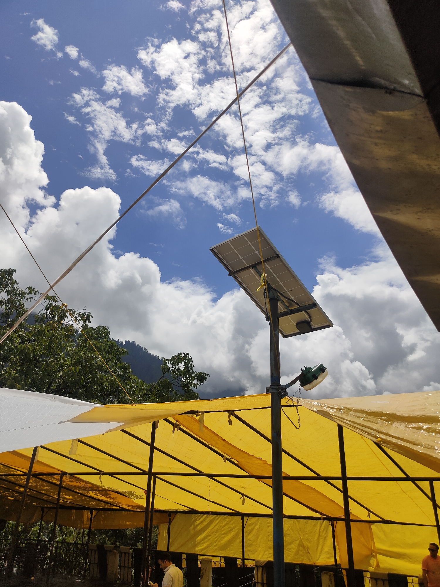 The image depicts an outdoor scene with a blue sky dotted with white clouds. In the foreground, a yellow canopy supported by a metal frame covers a structure. A solar panel is mounted on a pole above the canopy, with a light fixture attached to the pole. Trees and a mountain are visible in the background. The location is identified as Siyal, India. A man in a light-colored shirt is partially visible beneath the canopy, and another person wearing an orange shirt is on the right side of the image.