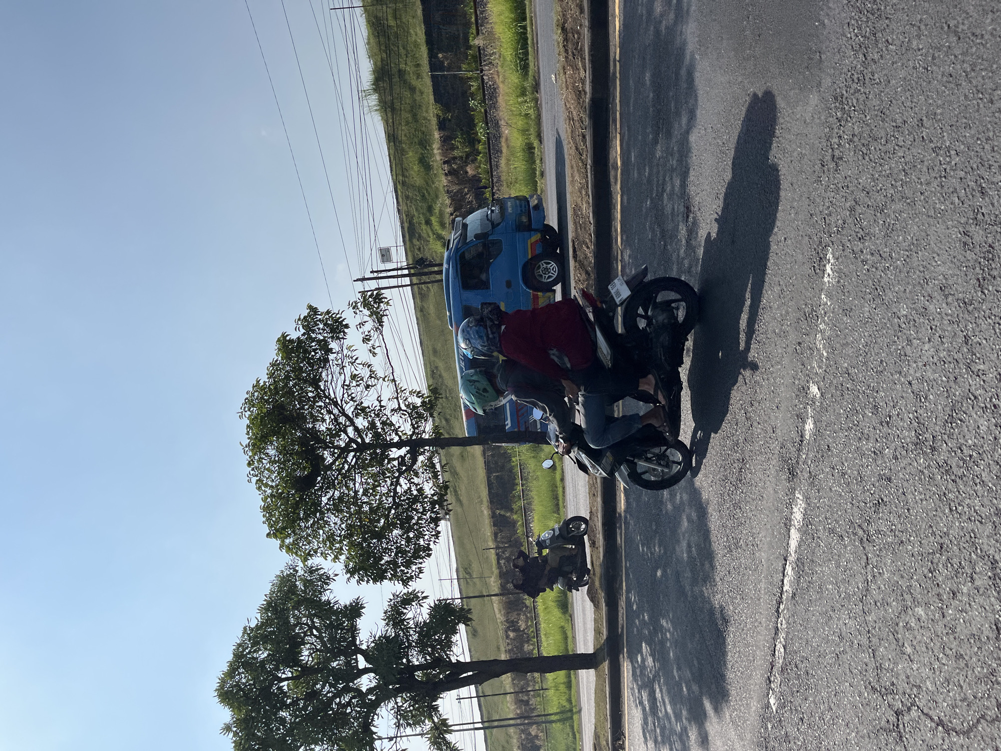 Two people are riding a motorcycle on a paved road in Pamotan, Indonesia, under a clear blue sky. The motorcycle is dark colored and the riders are wearing helmets. One rider is in front, wearing a green helmet, and the other is behind, wearing a blue helmet and a red jacket. A blue bus is visible driving on the road behind them, and to the right of the road, there is a grassy embankment with train tracks. Trees are on the left side of the road. The scene captures a typical moment of transportation in the Indonesian city.