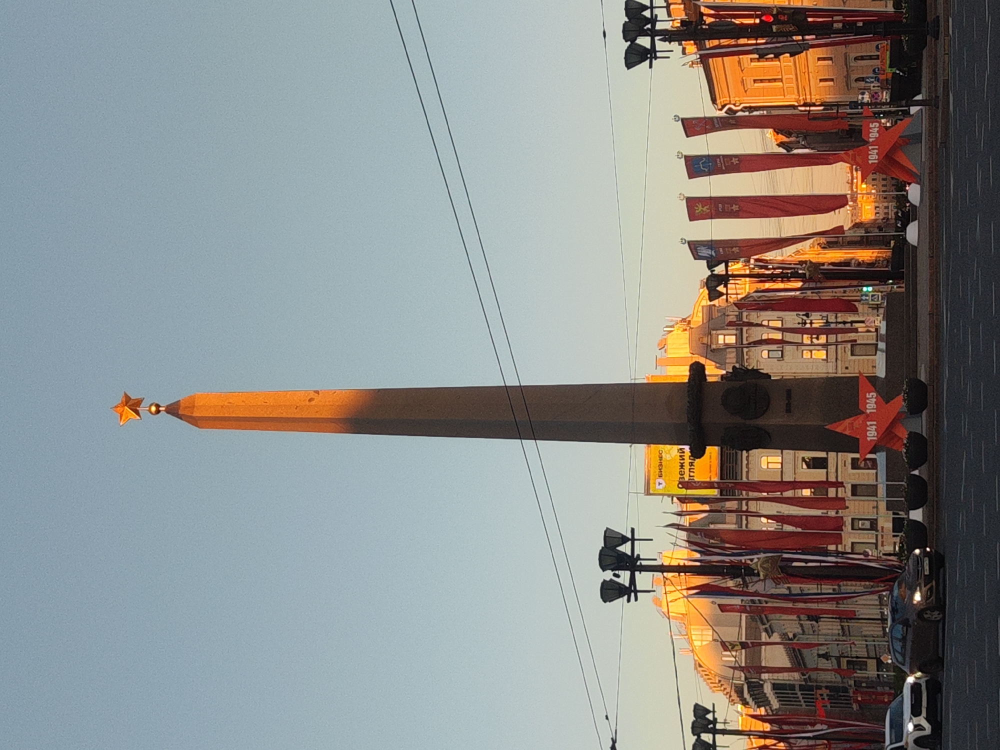 The image depicts the Alexander Column in Palace Square, Saint Petersburg, Russia. The column, topped with a star, is the central focus, with a clear sky behind it. Red flags, likely commemorating Victory Day, line the street on either side. Streetlights and buildings are visible in the background. Several cars are parked along the side of the road.