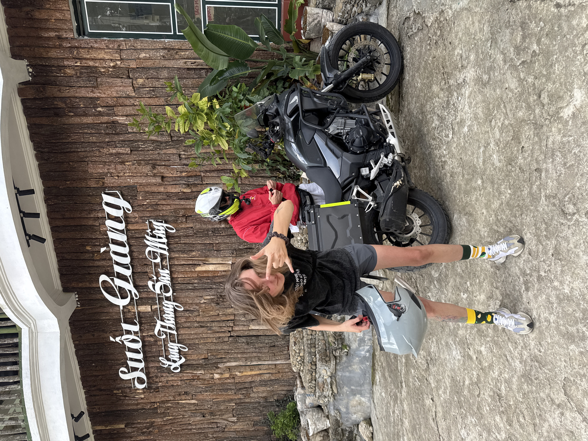 A daytime exterior shot shows two people, a motorcycle, and landscaping outside a building in Suối Giàng, Vietnam. A young, light-skinned person with long, blonde hair wearing a black t-shirt and shorts stands in the foreground holding a gray helmet, making a hand gesture, and looking at the camera. A gray motorcycle is positioned at an angle behind them, parked between the person and a person seated on the motorcycle. The seated person wears a red jacket and a white helmet. Behind the motorcycle and the seated person is a section of a wooden wall with a window and some greenery. To the left of the main subjects is a building with a white architectural detail and white lettering reading “Suối Giàng/Lung Thung Sui Ming." The ground is a rough, light-colored concrete or stone surface.