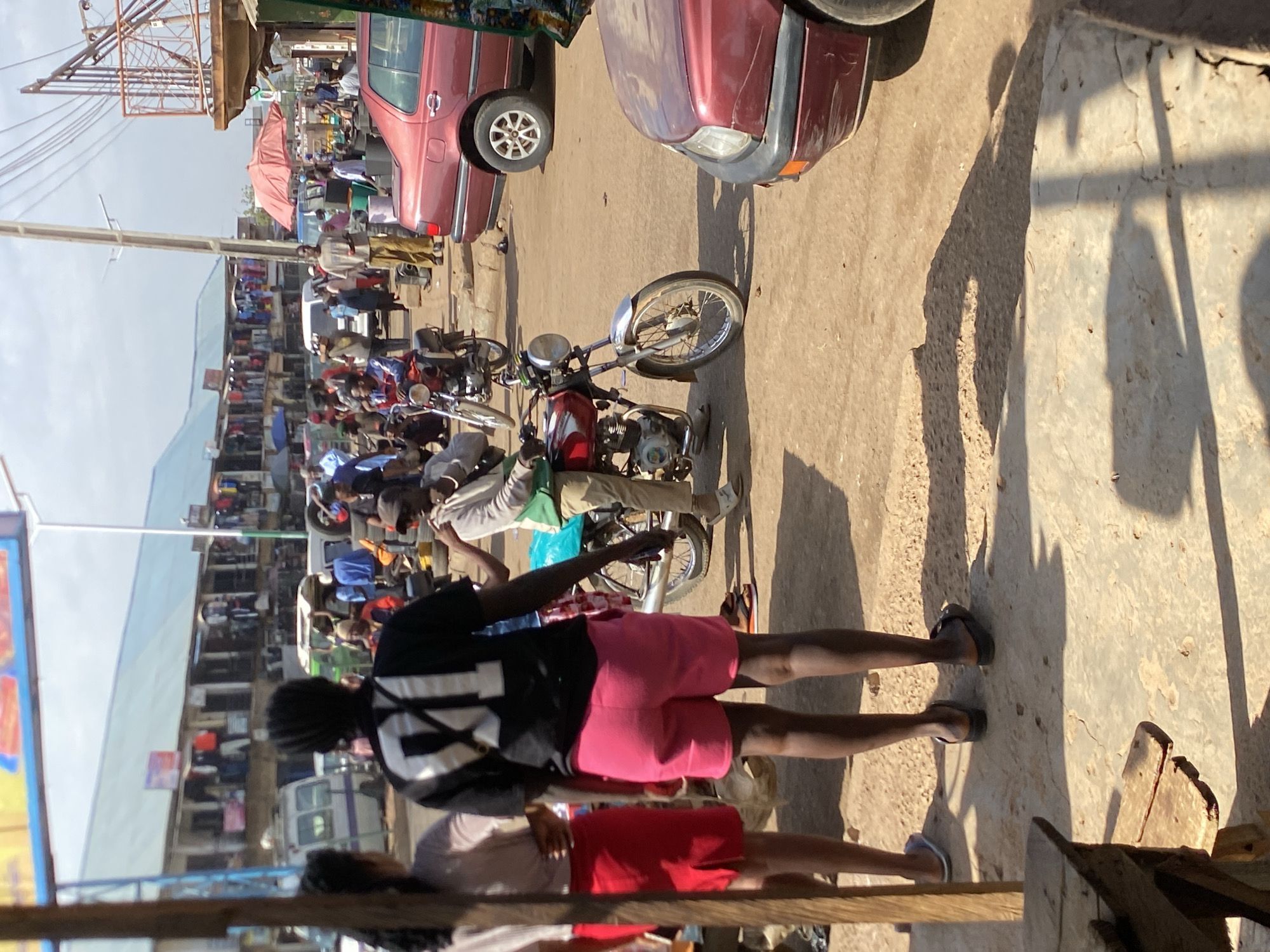 The image depicts a street scene in Bukuru, Nigeria. A large crowd of people is visible in the background. Several red cars and motorcycles are parked on the side of the road. In the foreground, two individuals are standing on the sidewalk. One person is wearing a black and white jersey and pink shorts. The other person is wearing a beige shirt and red shorts. The scene is sunlit.