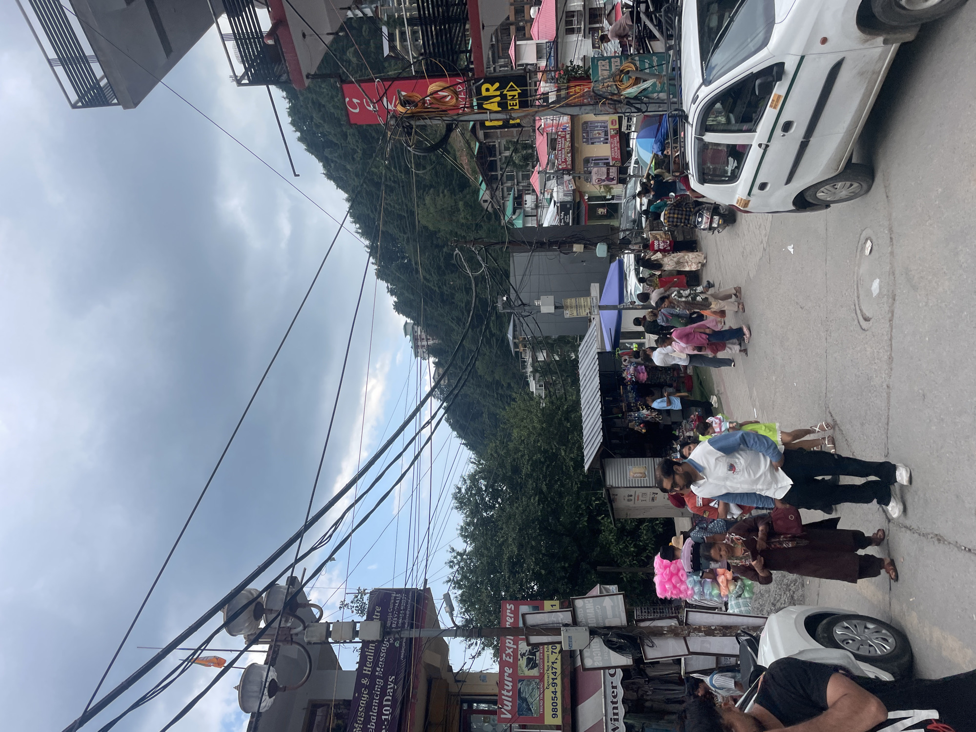 The image depicts a lively street scene in Bhagsu, India, under an overcast sky, with a lush, forested mountain serving as a prominent backdrop. Numerous overhead power and communication lines crisscross the frame, connecting to poles and the multi-story buildings that line the street.
The street is bustling with activity, showing many pedestrians walking, congregating, and browsing at various roadside stalls and shops. In the foreground, people are visible near displays of goods, including brightly colored items, suggesting a market or shopping area. A white car is parked on the right side of the street, and other vehicles, including a motorcycle, are present further down.
Notable details include the dense network of utility wires, the close proximity of buildings, and the vibrant street life. Visible text on various signs includes "Massage & Healing -10 Days balancing Massage" with contact information (82197-94415, 0905193937), and "Vulture Explorers" advertising "EKKING SCOOTY ON REN" along with phone numbers (98054-91471). Other partial signs include "AYURVEDA" and "Winter C." A red sign with a large white 'S' or '5' is also seen. The overall scene conveys a busy daytime atmosphere in a commercial area, with a blend of local businesses and a mountainous natural environment.