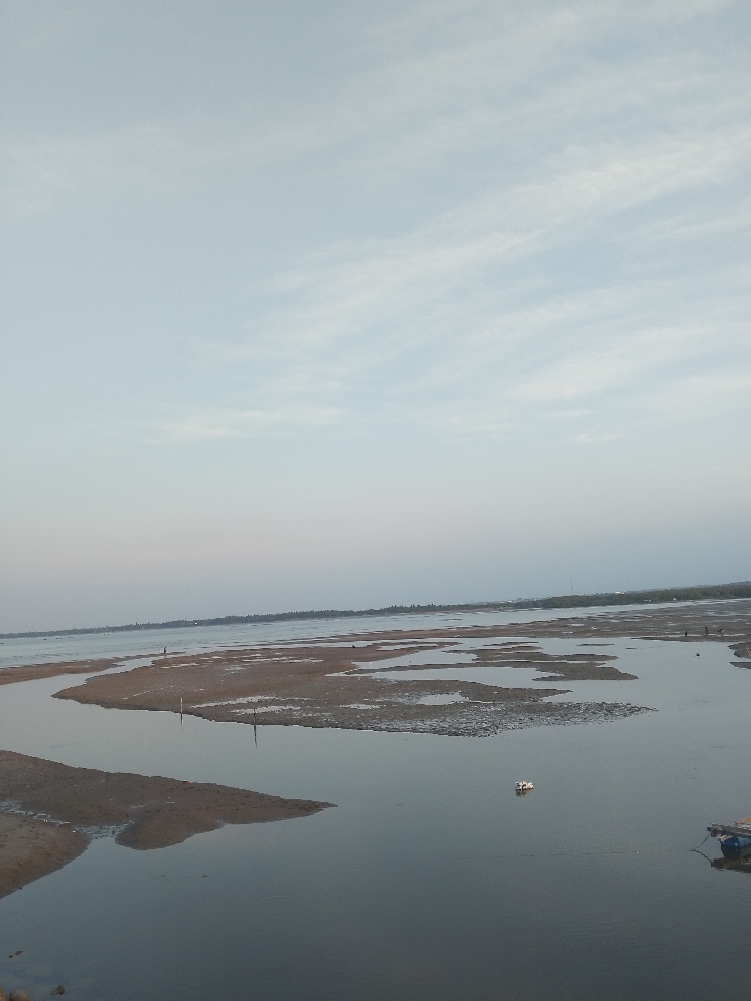 The image depicts a coastal scene in Lhokseumawe, Indonesia, at low tide. The foreground consists of a large expanse of muddy tidal flats interspersed with shallow water pools. Several figures are walking on the tidal flats in the mid-ground, appearing as small, indistinct silhouettes. In the distance, a thin strip of land lined with vegetation is visible, marking the coastline. The sky is overcast with light, wispy clouds, creating a soft, diffused light across the scene. A small boat is moored on the edge of the water, and further out, a cluster of floating debris can be observed. The overall impression is one of a serene, natural environment affected by tidal changes.