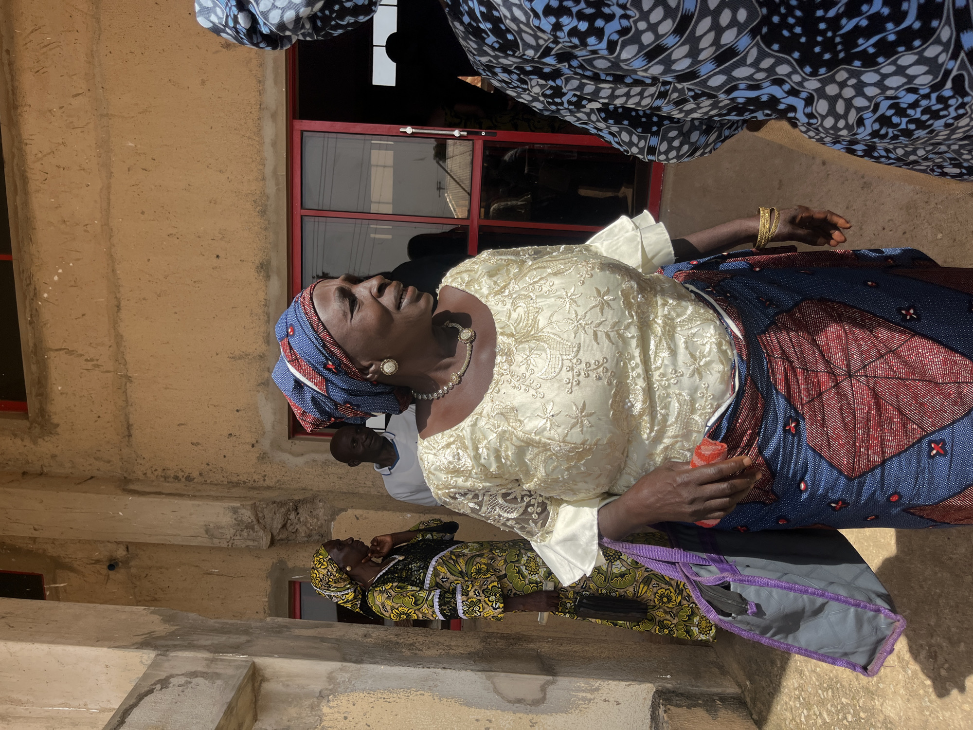 In Bukuru, Nigeria, an outdoor scene depicts several individuals gathered near a building entrance. In the foreground, a woman wearing a cream-colored lace top with ruffled sleeves and a vibrant blue and red patterned skirt is the central figure. She has a gold necklace and earrings, and a gold bracelet on her right wrist. She is holding a red cloth in her right hand and carrying a grey tote bag with purple trim in her left. To her left, another woman is visible, wearing a long green and yellow patterned dress and a yellow headscarf. She is holding a black object and a purple tote bag. Behind these women, a man in a white shirt is partially visible. In the background, a building with a tan-colored textured wall and red-framed windows is present. The lighting suggests it is daytime, with shadows cast on the ground. The overall atmosphere appears to be one of social gathering or event.