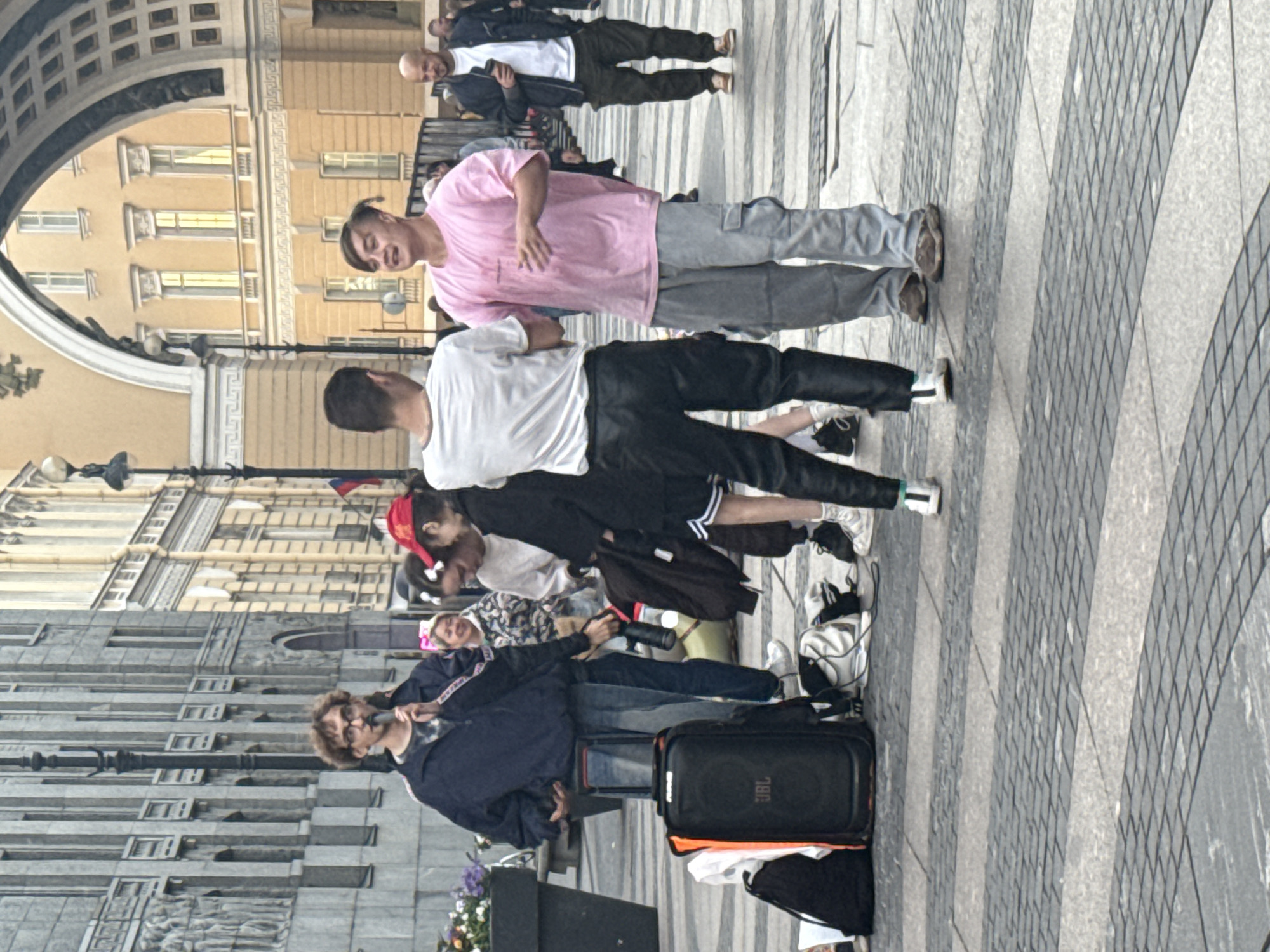 A street scene in Saint Petersburg, Russia, features a group of people gathered on a patterned cobblestone plaza. In the foreground, a man with glasses and curly hair speaks into a microphone, wearing a dark blue jacket and jeans. Beside him is a large black JBL speaker on wheels, along with bags and other equipment. To his right, two young men are conversing. One wears a white t-shirt and black pants, while the other sports a pink t-shirt and grey cargo pants. Behind them, several other individuals are visible, including a woman in a patterned jacket and a person wearing a red cap. In the background, grand neoclassical buildings with arched structures suggest a significant urban landmark, possibly part of the Palace Square or Nevsky Prospekt area. The architecture includes columns, arched windows, and ornate detailing. The overall atmosphere appears to be an outdoor event or gathering.