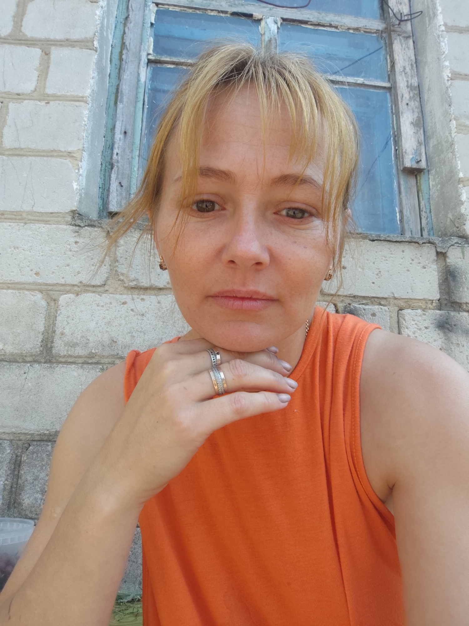 A medium shot shows a fair-skinned woman with blonde hair tied back and bangs, looking directly at the camera with a neutral expression. She is wearing a sleeveless orange top and has her left hand on her chin, displaying two rings on her fingers. She has on earrings and a delicate necklace. She is positioned in front of a light-colored brick wall with a weathered, blue-framed window behind her. The setting appears to be outdoors, possibly in the city of Saksahan, Ukraine.
