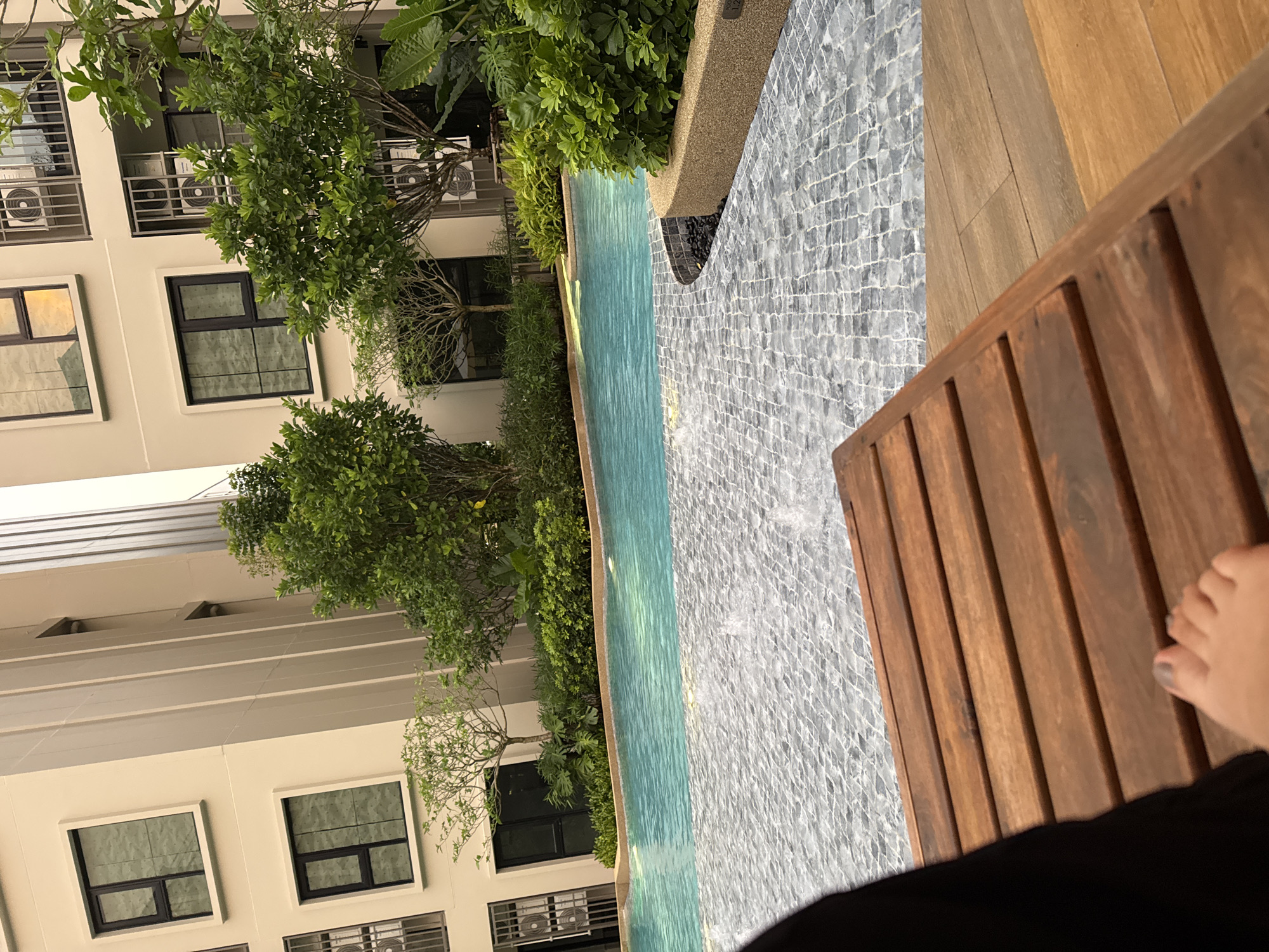 In Kathu, Thailand, a person's foot, with grey-painted toenails, rests on a wooden lounger in the foreground. Beyond the lounger, a swimming pool with turquoise water is visible, bordered by a stone-tiled walkway. Lush green vegetation and a modern building with windows and air conditioning units surround the pool area. The scene appears to be an outdoor common area within a residential complex.