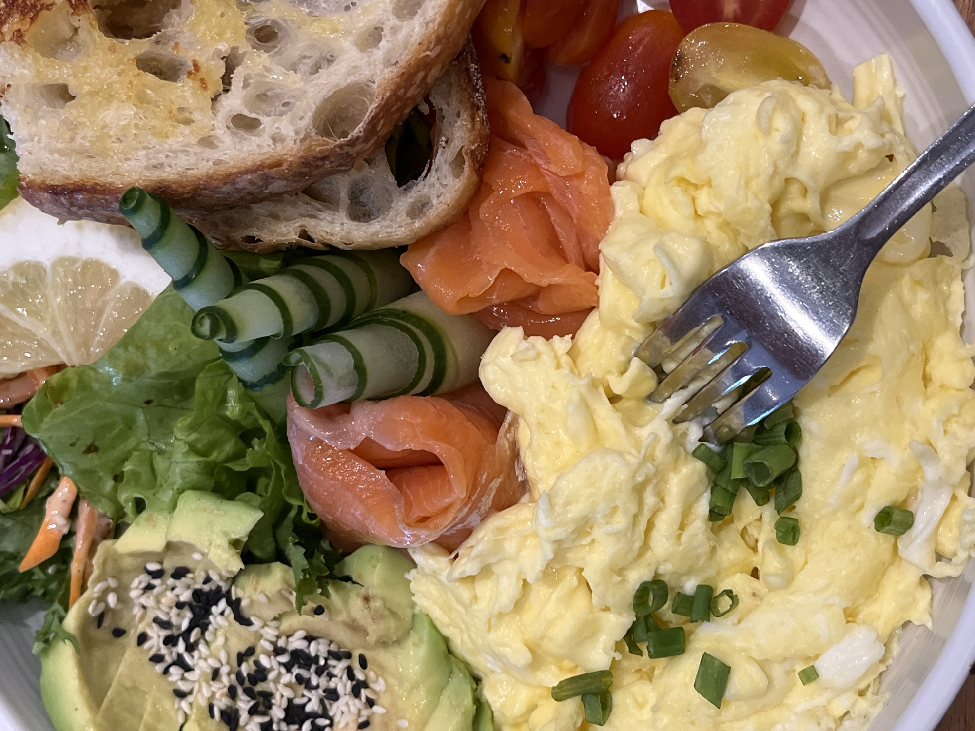 A close-up, overhead shot depicts a plate of food, likely a breakfast or brunch dish. The elements on the plate are arranged in a somewhat sectioned manner.
At the top left, there is a piece of toasted bread with large pores. Adjacent to it, there are slices of salmon. Below the salmon, there are spiralized cucumber slices and a section of lettuce with shredded carrots and purple cabbage.
On the bottom left, there is sliced avocado sprinkled with white and black sesame seeds.
To the right side of the salmon and avocado, there are scrambled eggs topped with chopped green onions. A fork is piercing into the scrambled eggs. A few cherry tomatoes are scattered among the food.
The image does not contain enough information to determine its location in Kuta Selatan, Indonesia. The scene depicts food arranged on a plate with a fork.