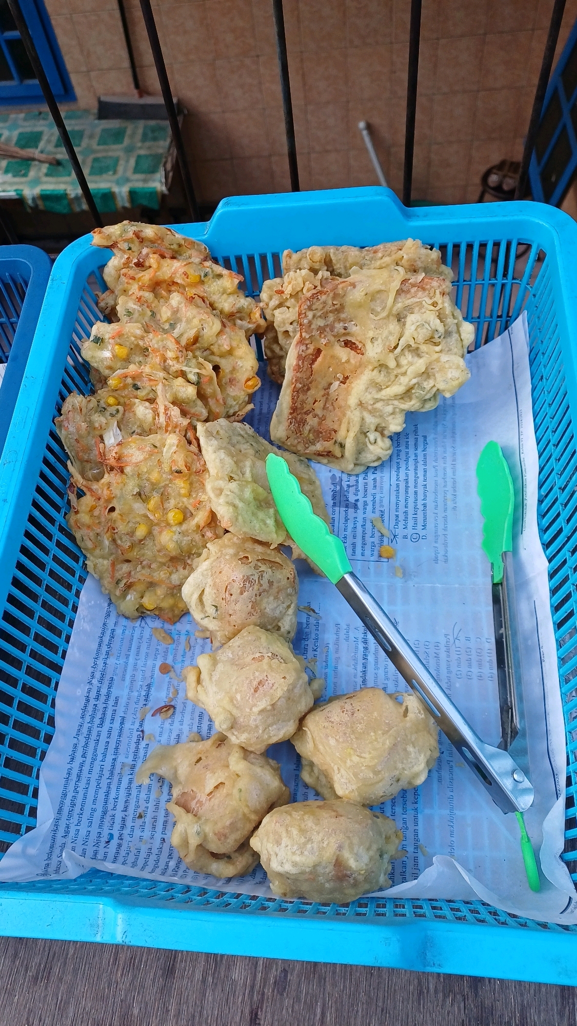 In Gunung Putri, Indonesia, an assortment of fried food items rests within a bright blue plastic basket. The basket is lined with white paper, some sections of which are covered in Indonesian text. At the top left of the basket, several pieces of fritters, likely bakwan sayur (vegetable fritters) due to visible corn kernels and shredded vegetables, are piled. To the right of these, larger, rectangular pieces of fried food, possibly tempe goreng (fried tempeh), are arranged. In the lower part of the basket, several round, golden-brown fried tofu or vegetable balls are visible. Two green-handled metal tongs are placed on top of the food, one resting across a piece of fried tempeh, and the other positioned near the fried tofu. The background shows a building facade with a tiled pattern and a blue window frame, suggesting an outdoor setting, possibly a food stall. The overall scene captures a selection of Indonesian street food, ready for serving.