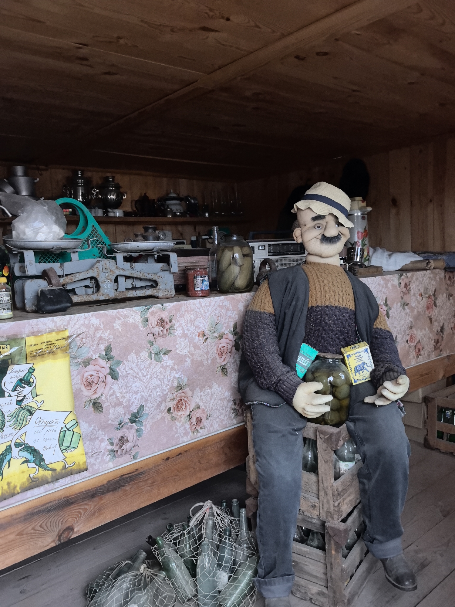 Here's a detailed description of the image:
**Location:** The image appears to be taken inside a small wooden structure, potentially a shop or storage area, located in the city of Bol'shoi Mashlyak, Russia.
**Subjects and Objects:**
* **Mannequin:** The focal point is a mannequin depicting a man. He is wearing a straw hat, a sweater, dark trousers, and shoes. He sits on a stack of wooden crates. The mannequin is holding a jar of what appear to be pickled vegetables (possibly cucumbers). Labels are attached to the mannequin, including one with the word "чай" which translates to "tea."
* **Pickled Goods:** Jars of pickled items are also visible on a table behind the mannequin.
* **Scales:** An old, rusty weighing scale sits on the table behind the mannequin.
* **Bottles:** On the floor to the left of the mannequin are several green bottles, some bundled in netting.
* **Table:** A table with a floral-patterned covering runs along the back wall.
* **Shelves:** Above the table, shelves are filled with various items, including pots, pans, teapots, and other containers.
* **Poster:** A poster with illustrations and text is visible on the left side of the table. The poster includes the words "Устрой" and "Gпусти."
**Actions and Interactions:**
* The mannequin is posed to appear as if he's sitting and holding a jar of pickles.
* The overall scene suggests a store that sells or displays pickled vegetables and other goods.
**Environment:**
* The interior is enclosed with wooden walls and a wooden roof.
* The lighting is somewhat dim, suggesting either indoor artificial lighting or daylight coming in from a limited source.
* The atmosphere feels somewhat rustic and old-fashioned.