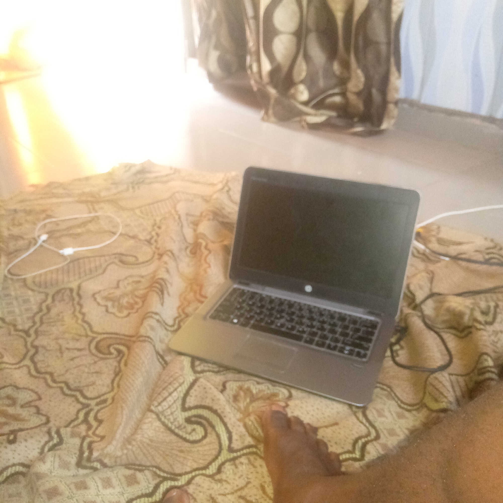 An overhead, slightly blurry shot captures a laptop resting on a patterned blanket. The blanket features a repeating motif of swirling lines and diamond shapes in brown and yellow. A pair of white earbuds is also visible on the blanket, loosely coiled near the laptop. To the right of the laptop, a person's bare foot, with dark skin and painted toenails, is visible. The foot appears to be resting on the blanket as well. In the background, a curtain with a similar patterned design hangs, and sunlight streams in from the left, creating a bright, washed-out effect. The overall impression is one of a casual, indoor setting.