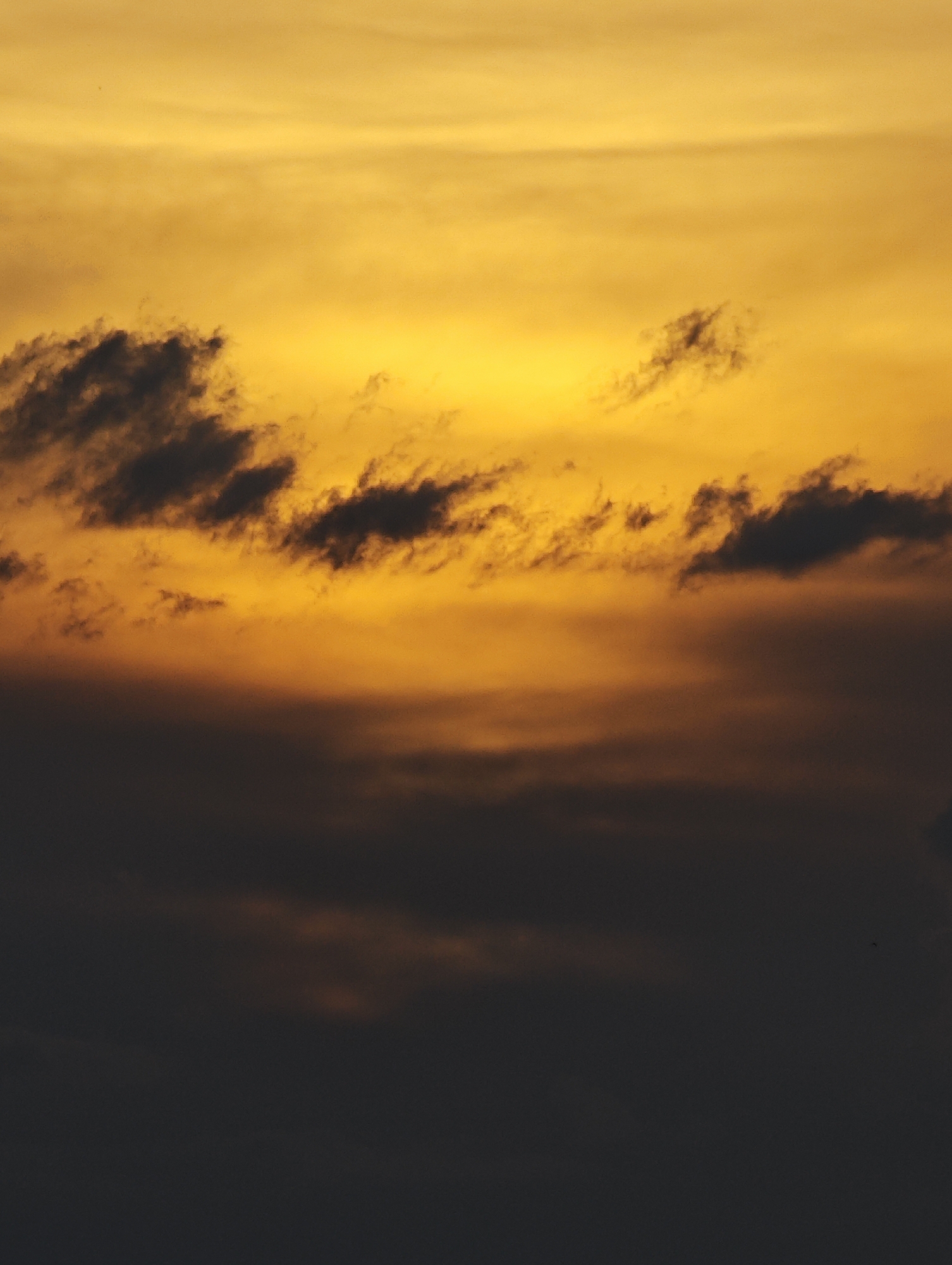 The image depicts a dramatic sky with a gradient of colors ranging from bright yellow at the top to dark grey at the bottom. Scattered clouds are visible, some dark and dense, others appearing as wisps against the bright sky. The overall impression is that of a sunset or sunrise. The scene is an outdoor environment, likely the sky above a city. No specific landmarks or elements that would definitively place it in Sofia, Bulgaria, are present in the image. The image is devoid of people, subjects, objects, or any discernible actions or interactions.
