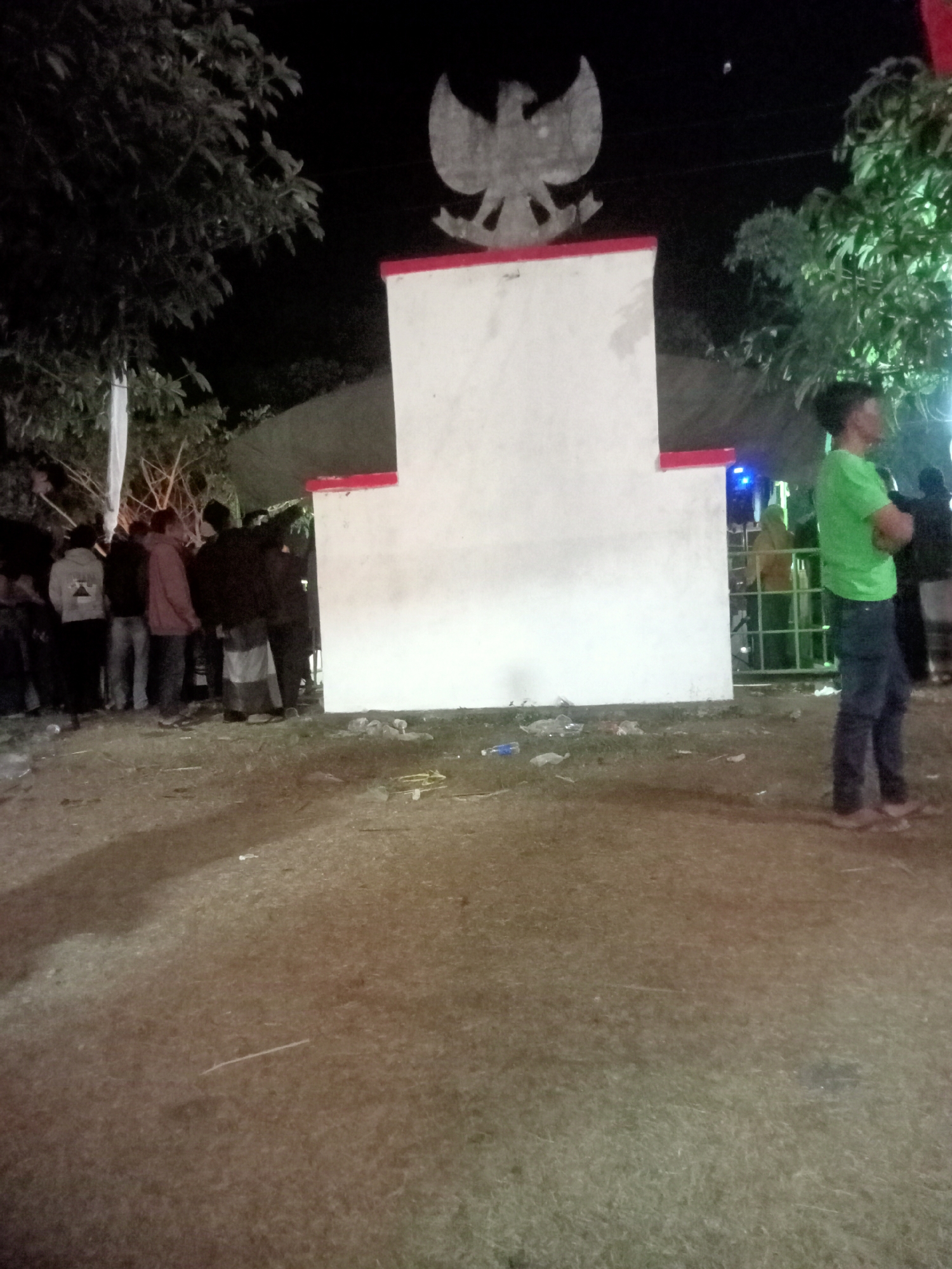 In Pule, Indonesia, at night, a monument shaped like a large white rectangular pillar with a Garuda symbol at its apex stands in an outdoor area. The monument has red trim along its top edges. Several individuals are present around the monument. To the left of the monument, a group of people are gathered, some wearing casual clothing. To the right, a person in a green t-shirt and blue jeans is standing with arms crossed, facing away from the camera. In the background, there appears to be a tent or canopy, and some trees are visible. The ground is unpaved, with scattered debris and shadows indicating artificial lighting.