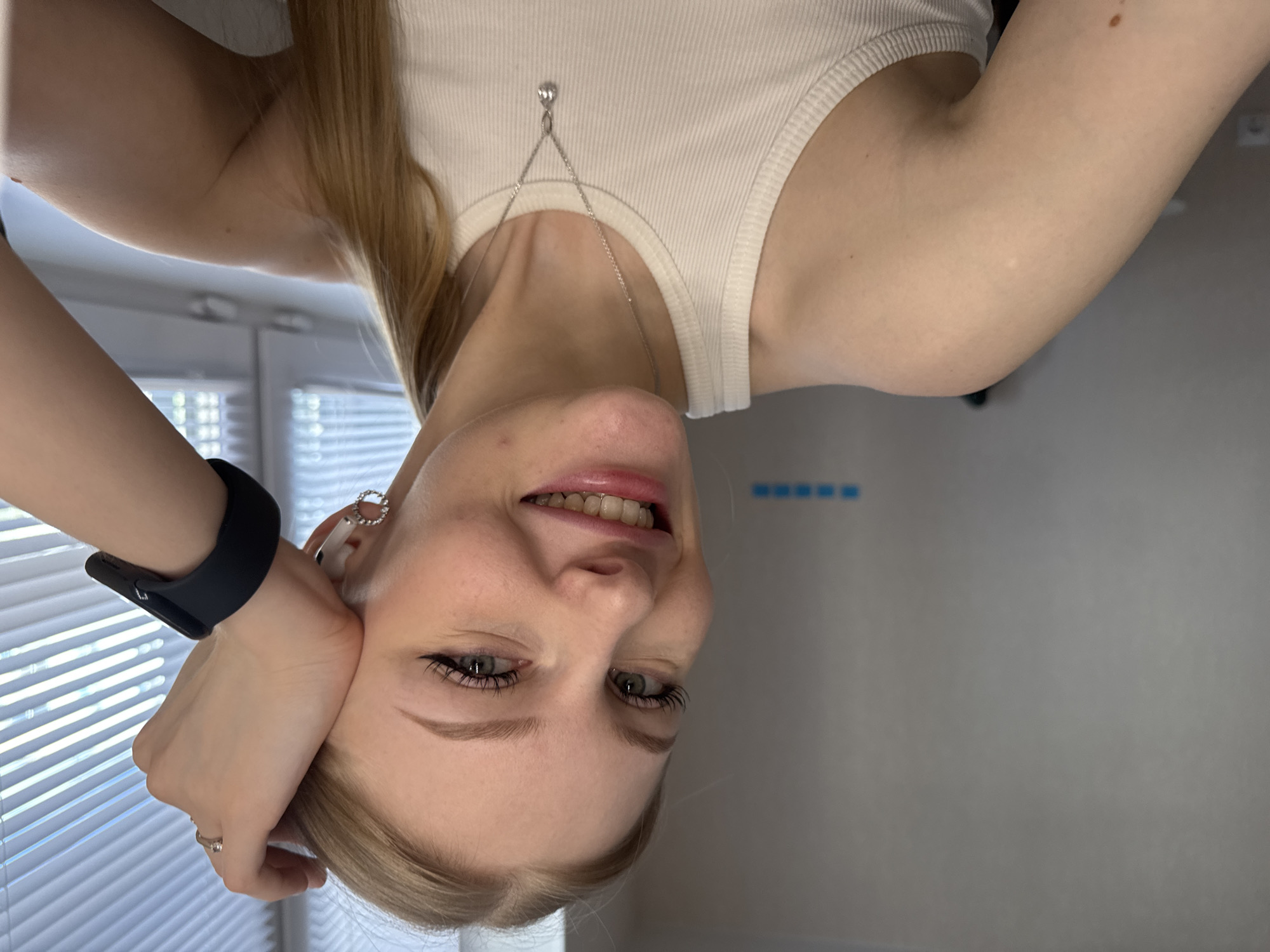 A woman is seen from an inverted perspective, with her face angled toward the camera. She is wearing a white tank top and a silver necklace. Her left hand is positioned near her face, adorned with a black smartwatch and a ring. She has blond hair, and her eyes are visible. The background features white window blinds. The location, Perm, Russia, is given.