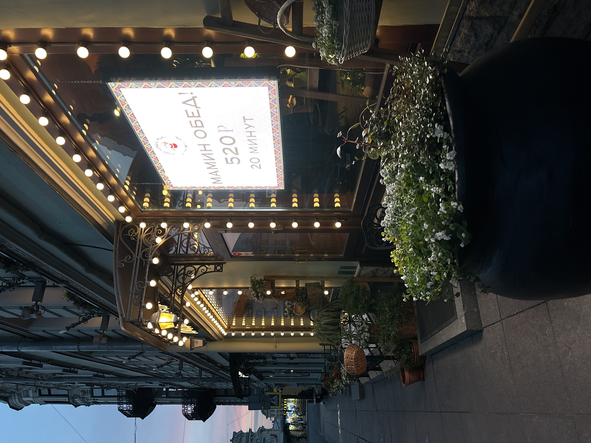 The image depicts the facade of a building in what appears to be Saint Petersburg, Russia. The building is illuminated by multiple rows of small, round lights. A sign above the entrance advertises "MAMIN OBED!" (Mother's Lunch) at a price of 520 rubles and a preparation time of 20 minutes. The entrance is decorated with plants and floral arrangements, including baskets and potted plants. The sidewalk in front of the building is visible, and the street stretches into the distance, with other buildings visible on both sides. The sky in the distance has a pinkish hue, suggesting either dawn or dusk. Two ornate streetlamps hang from the buildings along the street.