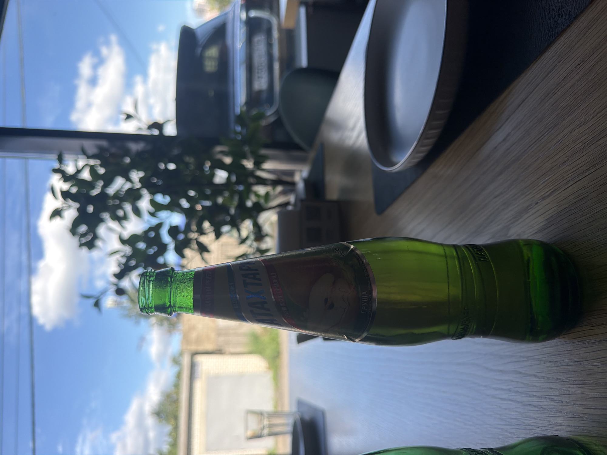 The image shows a green glass bottle of "АTAXTAP" brand pear-flavored Georgian lemonade, standing on a wooden table. The bottle has a label with Cyrillic text and an illustration of a pear. In the background, through a window, a blue sky with clouds and some foliage are visible. Outside, a white garage-like building and a dark-colored SUV with a license plate are discernible. The scene appears to be set in an outdoor cafe or restaurant, possibly in Kazan, Russia, given the context provided. The lighting suggests it is daytime. No people are visible in the image.