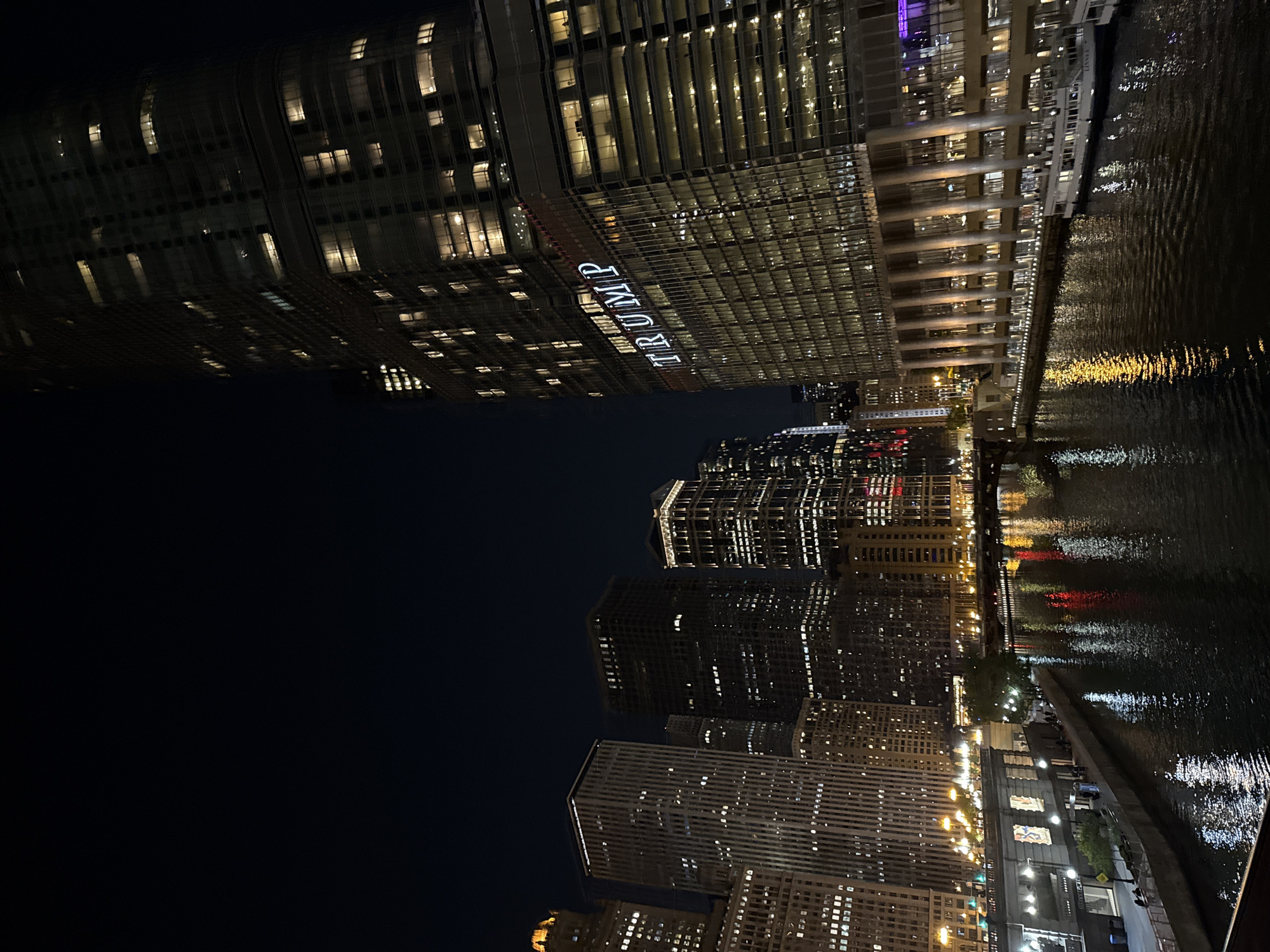 The image depicts an urban night scene in Chicago, United States of America, featuring the cityscape alongside a body of water, likely the Chicago River. Notable landmarks include the Trump International Hotel and Tower, illuminated with the word "TRUMP" prominently displayed on its facade. Other high-rise buildings, glowing with interior lights, line the waterway. The river's surface reflects the city's lights, creating a shimmering effect. The scene is viewed from a low angle, offering a perspective that emphasizes the height and scale of the buildings. Along the edge of the river, there's a visible sidewalk or pathway.