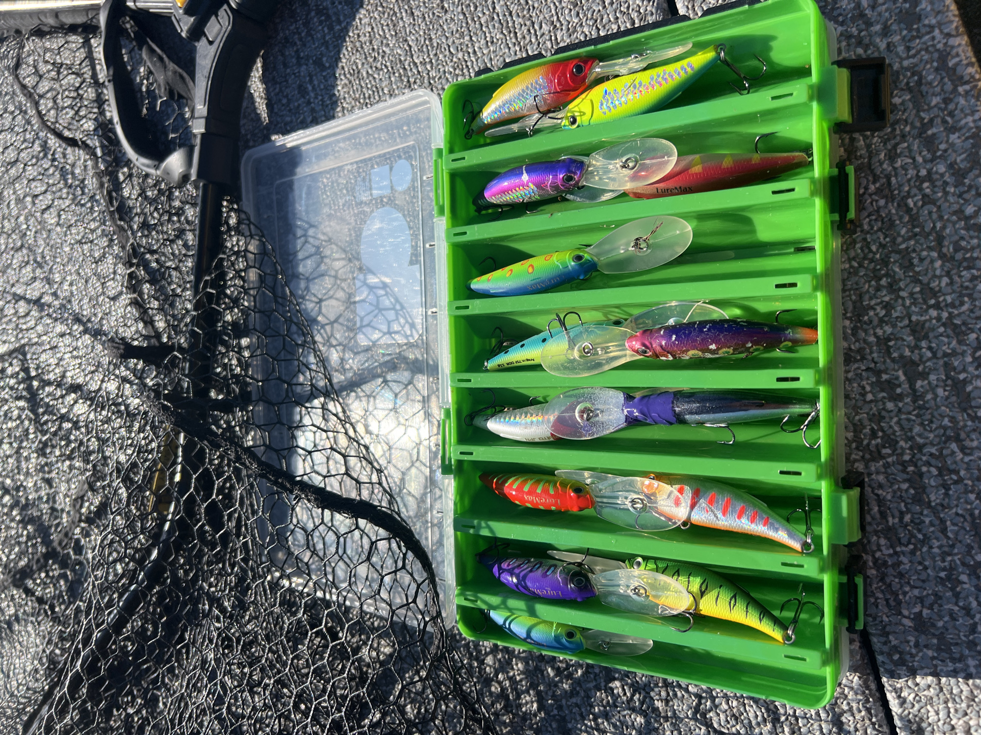 The image shows a bright green fishing tackle box filled with an assortment of colorful fishing lures. The tackle box is open, revealing multiple compartments, each holding several lures. The lures vary in color and design, featuring shades of red, yellow, blue, purple, and green, with some having reflective or iridescent finishes. Most of the lures are clearly designed to mimic fish or other aquatic prey.
Beside the tackle box, a black fishing net is visible, partially covering a clear plastic container that appears to be another storage item. The background is a textured, grey surface, likely the deck of a boat. Sunlight is casting strong shadows, suggesting it is a sunny day, possibly late morning or early afternoon. The scene appears to be set outdoors, likely on the water, given the fishing gear and the presence of a boat deck.
There is some visible text on the lures, including "LureMax" on at least two of them. Another lure has text that is partially obscured but appears to include "JAPAN." The overall impression is one of preparation for or a break during a fishing trip.