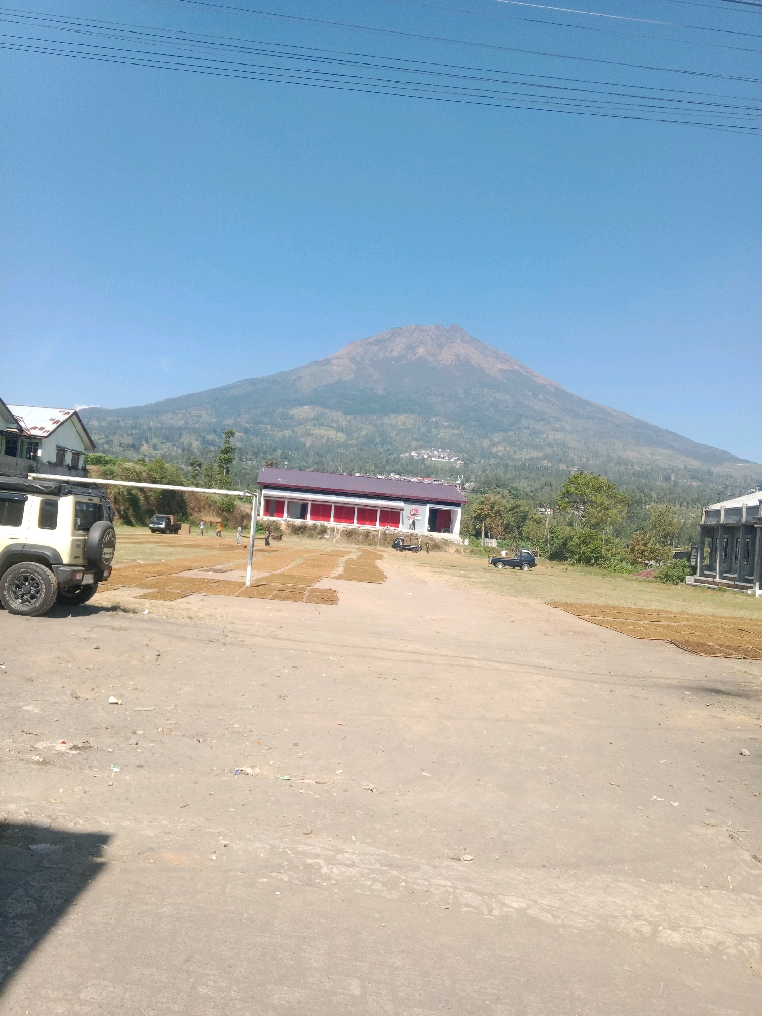 In Kapencar, Indonesia, an outdoor scene unfolds under a clear blue sky. In the foreground, a dirt and gravel lot is partially covered by a shadow on the left. Several vehicles are present, including a white Jeep with a spare tire mounted on the rear, positioned on the left side of the frame. Further back, a small black truck is visible, and another dark-colored pickup truck is parked in the middle ground.
The middle ground of the image features several buildings and an open grassy area. One building, with a dark roof and white walls, is on the left. Another building, with a red and dark-colored roof and white walls with red accents, is in the center. This building appears to be a commercial establishment. To the right, another building with a dark, modern facade and large windows is seen. Scattered across the grassy area in front of the buildings are numerous rectangular piles of what appear to be dried leaves or plant material, possibly for drying or processing. A volleyball net with white poles stands in the middle of the field. Several individuals are casually walking or standing in the vicinity of the buildings and the drying materials.
Dominating the background is a large, majestic mountain with a cone-shaped peak, its slopes covered in vegetation and dotted with small settlements. The mountain is a prominent natural feature in the landscape. The overall environment suggests a rural or semi-rural setting, likely associated with agricultural activities, given the presence of the drying materials and the proximity to the mountain.