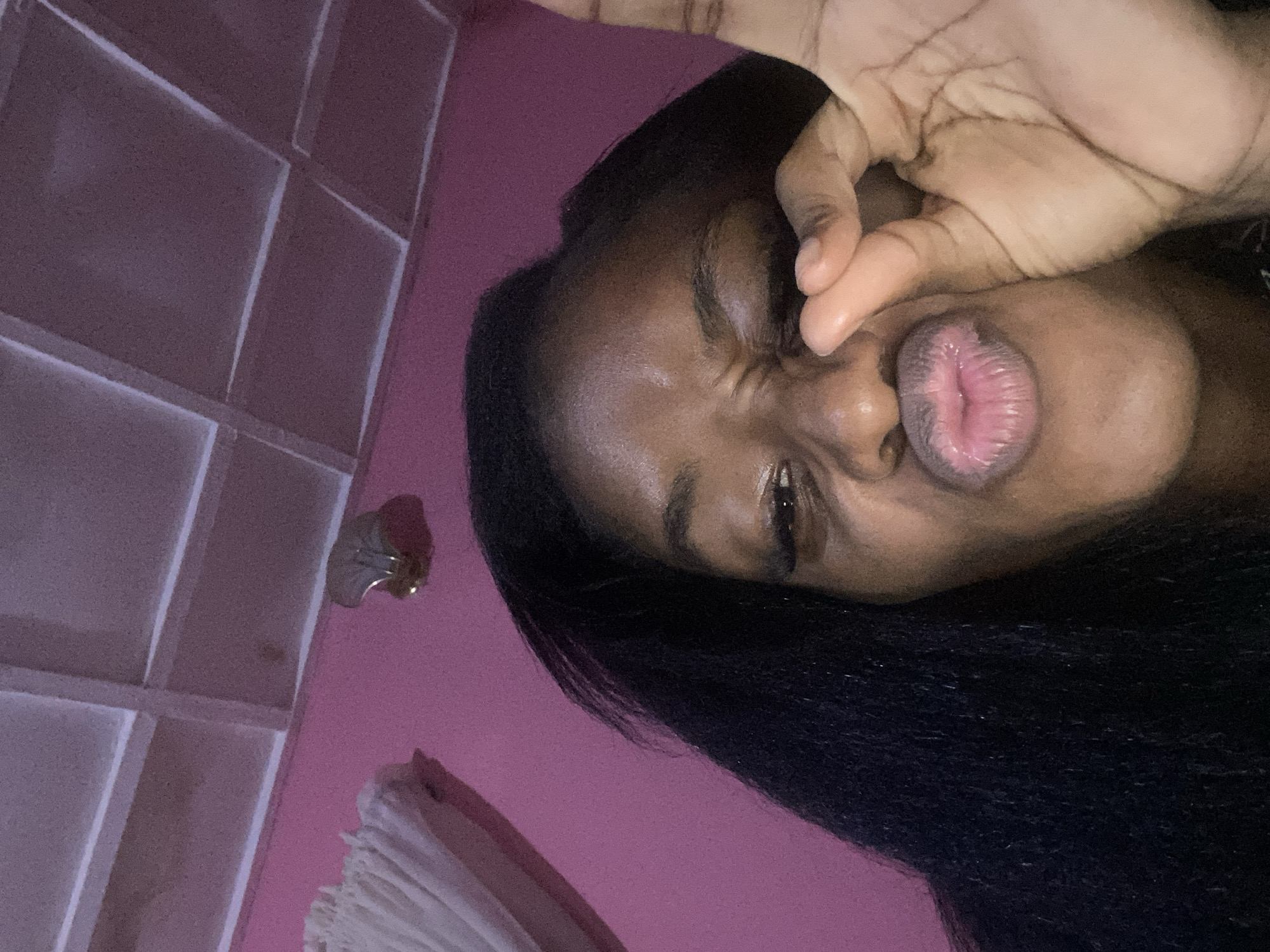 The image shows a close-up, low-angle selfie of a young Black woman with long, dark hair. She is making a "pucker" face with her lips and pinching her cheek with her right hand, forming an "okay" gesture with her thumb and index finger. Her left eye is squinted, and her right eye is partially visible. The background is a pink wall with a white, tiled ceiling above it. A section of white curtains is visible on the left side of the image. A metallic, shell-shaped object is attached to the pink wall near the top center of the frame. The lighting suggests an indoor setting, possibly a bedroom, given the pink decor. There are no identifiable landmarks or clues within the image that would indicate the specific location within Jos, Nigeria. The image appears to be taken in a domestic setting.