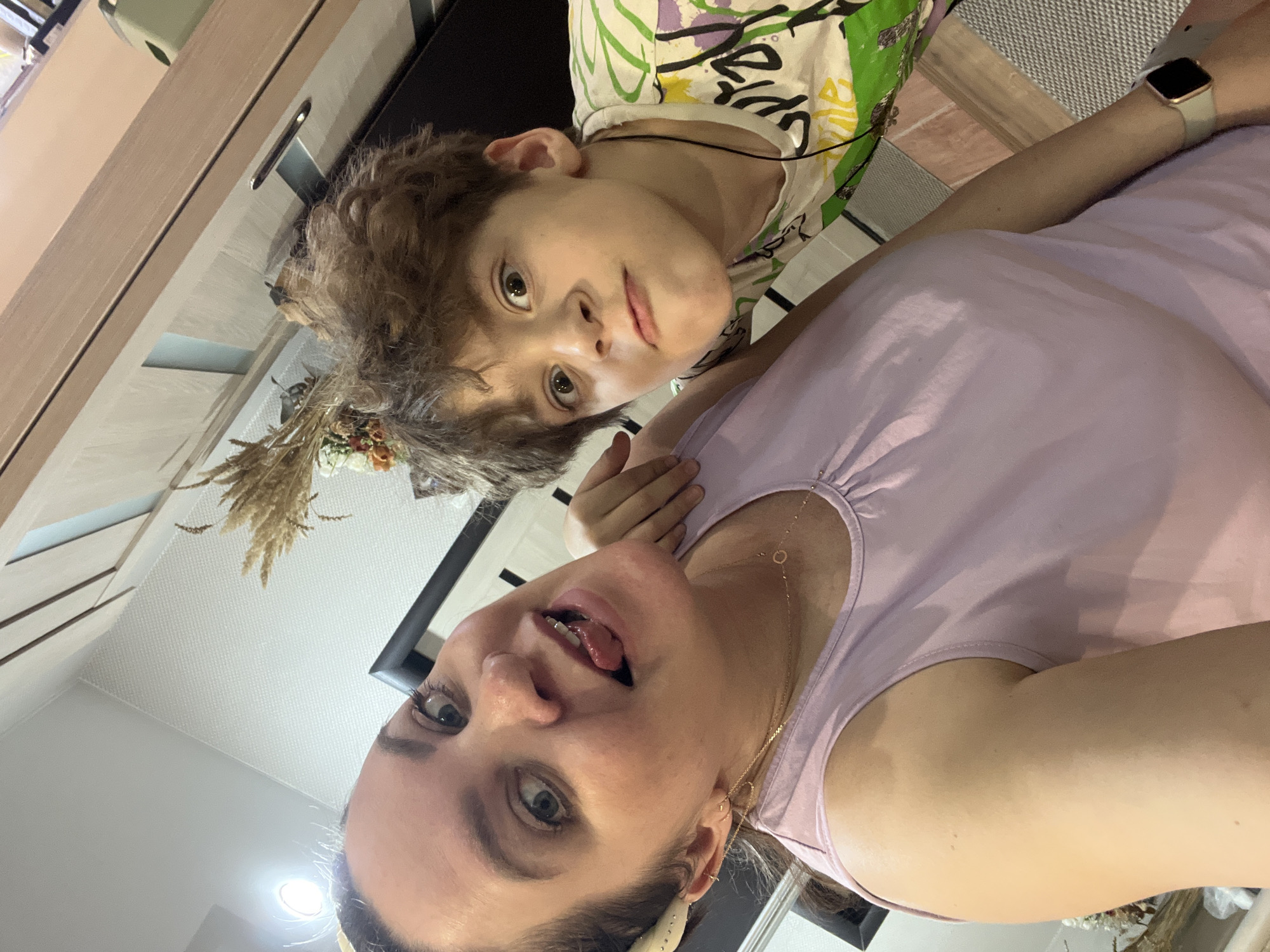 This image captures two individuals, an adult woman and a child or teenager, in an indoor setting, likely a home in Noviy, Russia. The photo appears to be a selfie, taken from a slightly upward and tilted perspective, suggesting a casual and intimate moment.
The woman, positioned in the foreground on the right, has fair skin and dark hair, which is pulled back. She wears a light purple sleeveless top and accessorizes with a delicate gold necklace and small gold earrings. Her expression is playful, characterized by wide eyes and her tongue slightly extended. A smartwatch with a light-colored band is visible on her left wrist.
Behind her and positioned more centrally is the child or teenager, who has fair skin and light brown, curly hair. They are dressed in a white t-shirt featuring a vibrant, abstract pattern with green, purple, yellow, and black elements. The child looks directly at the camera with a more serious or neutral expression. A decorative element, possibly a dried floral or wheat arrangement, is visible near the child's head, suggesting part of the room's decor.
The background reveals elements of a modern interior. On the left side, light-colored wooden cabinetry or shelving is visible, and above it, a light-colored ceiling with a recessed light fixture. To the right, behind the subjects, there is a dark surface, possibly a counter or furniture, and a textured rug or mat. No discernible text is visible within the image. The overall scene suggests a warm, informal atmosphere.