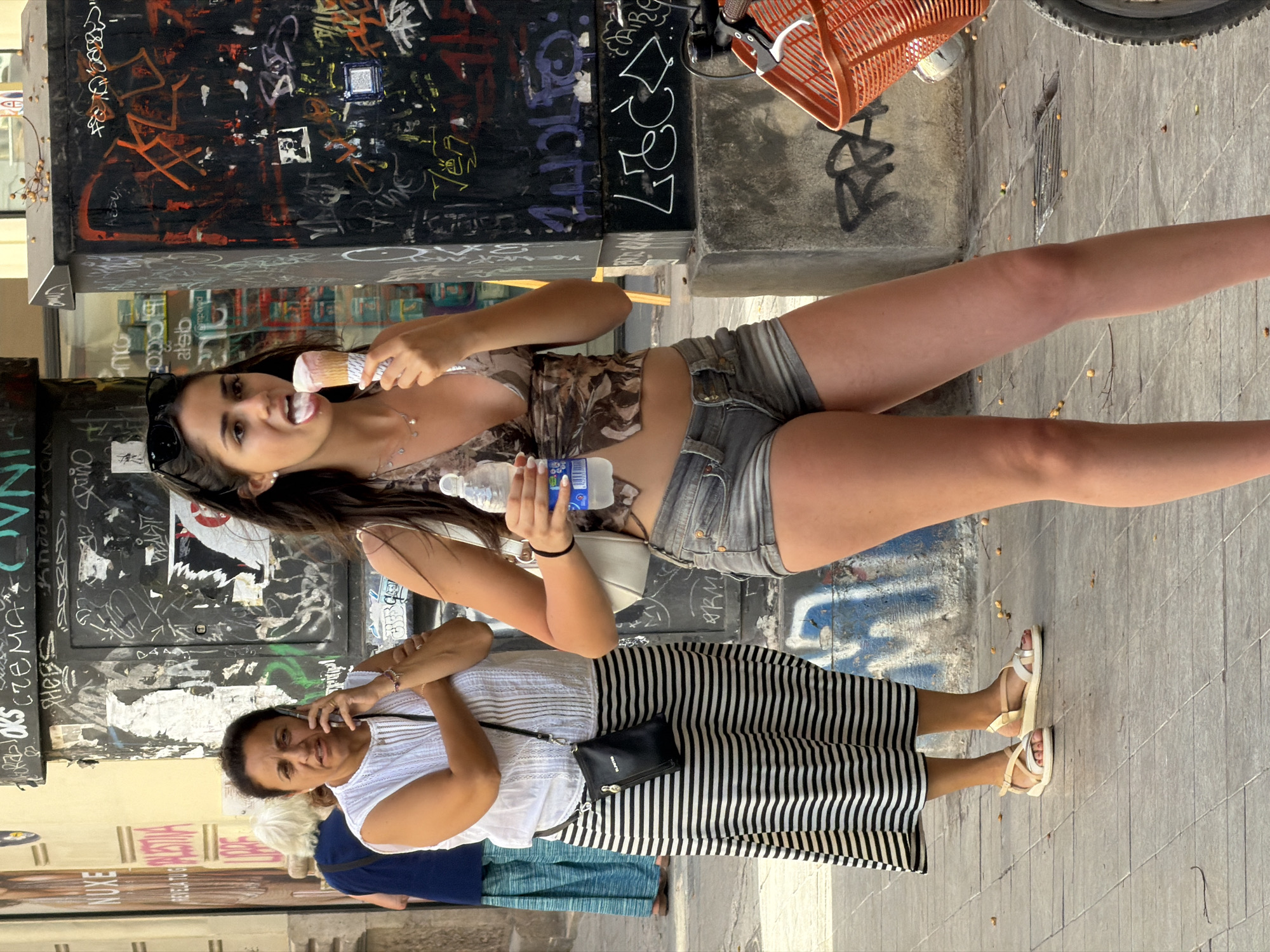 In an urban environment, possibly a city street, a young woman with dark hair and sunglasses perched on her head is captured mid-action, licking an ice cream cone. She wears a patterned top and denim shorts, holding a bottle of water in her left hand. To her left, an older woman in a white top and a black and white striped skirt speaks on her cell phone. In the background, a person with blond hair wearing a blue shirt and long blue skirt walks away from the camera. The scene is set against a backdrop of a building facade covered in graffiti, with a bicycle basket in the foreground. The ground is paved with grey tiles.