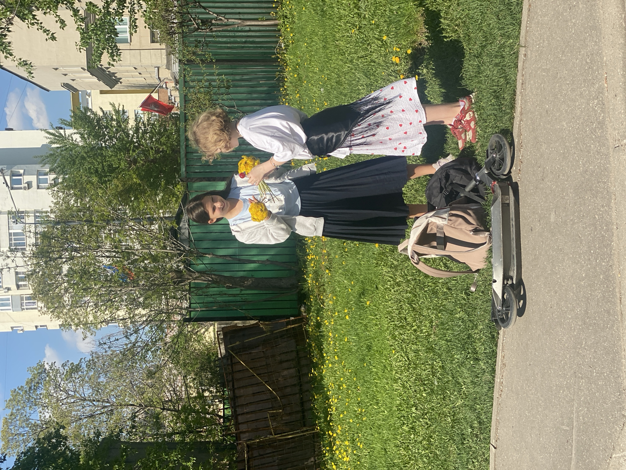 The image shows two girls standing on a grassy patch next to a sidewalk in a city environment, possibly Moscow based on the architecture and a flag visible in the background. The grass is dotted with yellow flowers, likely dandelions. A green metal fence is visible in the background. One girl is wearing a white shirt and a white skirt with red hearts, and red sandals. The other girl is wearing a light blue top, a white cardigan, and a dark blue pleated skirt. They both appear to be examining a bunch of yellow flowers. A scooter and two backpacks, one black and one tan, are resting near the sidewalk. Buildings are visible behind the trees, including a white building with multiple windows and what appears to be a red flag hanging from a building.