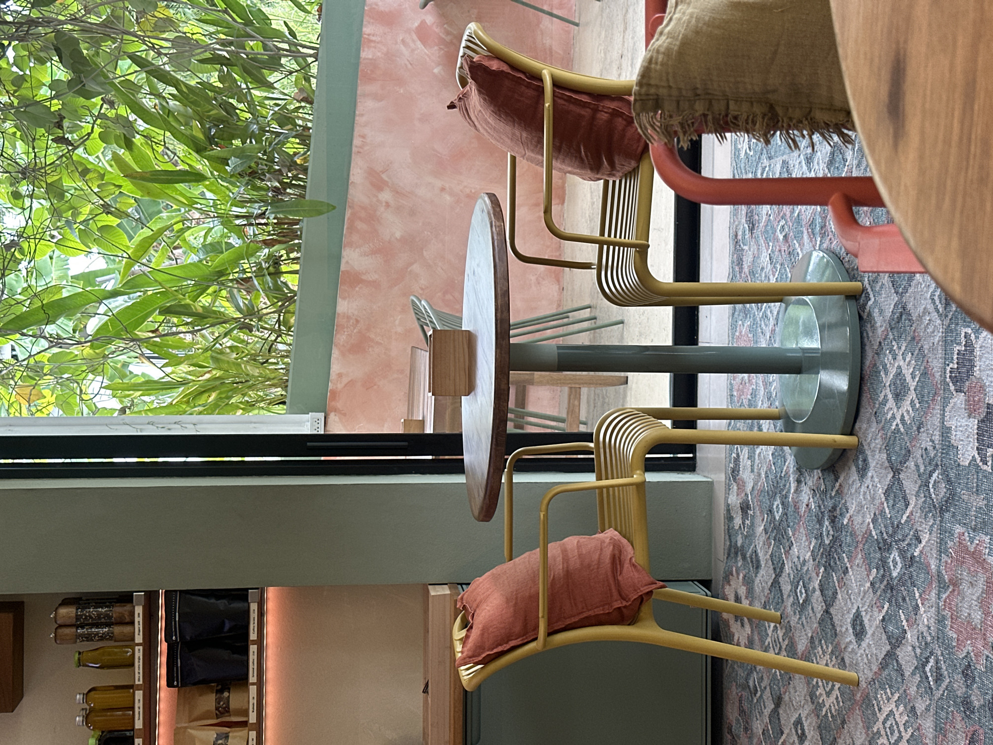 The image depicts a cafe interior with a focus on seating arrangements and decor. The scene is set during the daytime, with natural light filtering in from a large window covered by lush green foliage. The cafe appears to be located in Koh Phangan, Thailand, as indicated by the context.
The primary subjects are two chairs with yellow metal frames. One chair, in the foreground, has a textured, coral-colored cushion placed on its seat. Another similar chair, partially visible in the background, also has a cushion, this one appearing to be a muted tan color with fringe detail. Between the chairs, a round wooden table is supported by a light green pedestal base. The table has a wooden top and a green cylindrical leg.
The flooring is covered by a patterned rug with a diamond motif in shades of grey, pink, and blue. In the background, a wall painted in a soft, textured pink hue suggests a warm and inviting atmosphere. To the left of the frame, shelves are stocked with various items, including bottles of what appear to be sauces or beverages, and packages of coffee or other food items. Some of these packages have visible text, but it is too small and blurry to be legible.
The overall impression is one of a stylish and comfortable cafe, likely designed to offer a relaxed ambiance. The presence of the plants suggests a connection to nature, possibly an outdoor or garden-themed setting. No people are visible in the image, and the scene is still and quiet, with no apparent activity taking place.