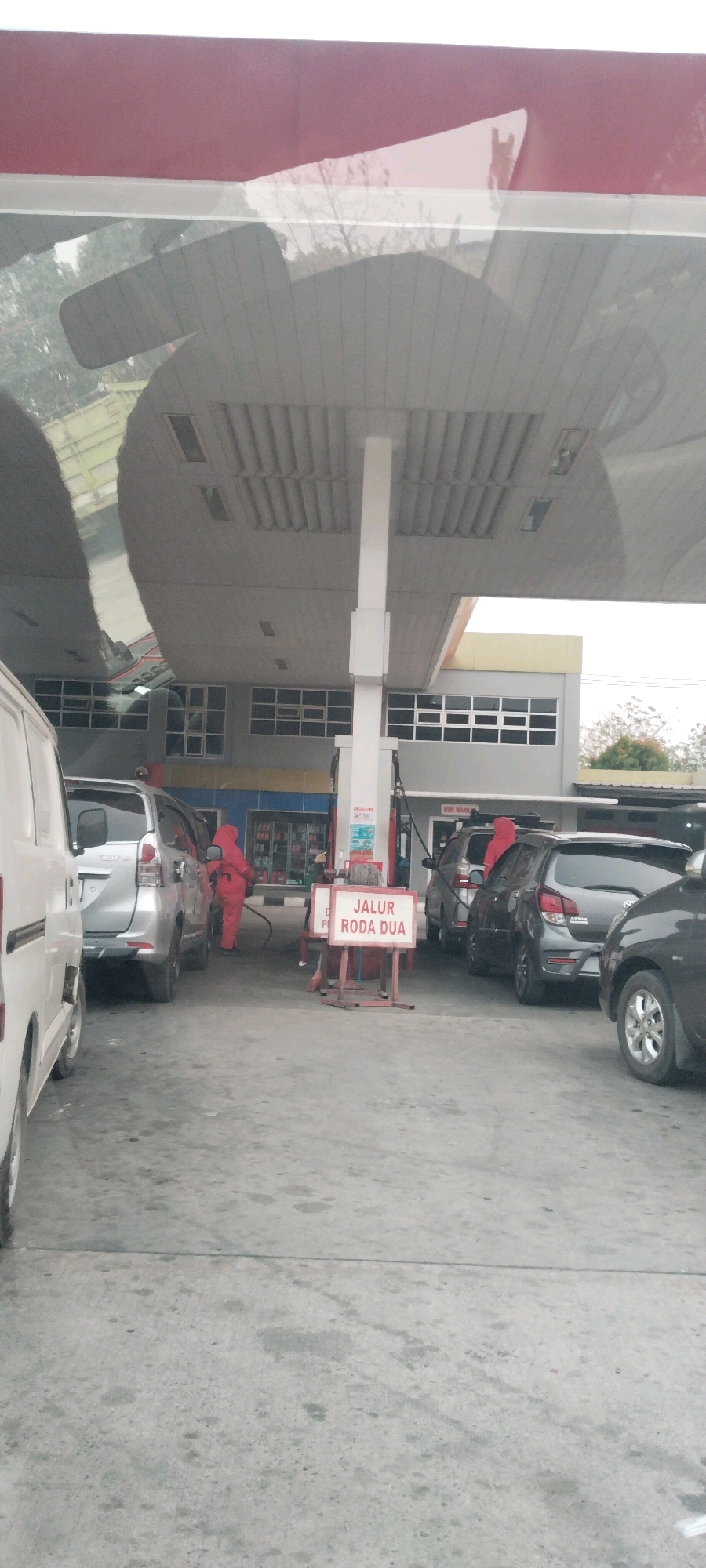 The image depicts a scene at a gas station in Haduyang, Indonesia. A person dressed in a bright red uniform, likely an attendant, is fueling a silver car. Several other vehicles, including a white van, a dark grey car, and another silver car, are present at the station. A sign with "JALUR RODA DUA" (two-wheel lane) is visible in the foreground, indicating a designated area. The gas station appears to be part of a larger complex, with a building housing what might be a convenience store in the background. The ground is paved, and the overall environment suggests a typical urban gas station setting.