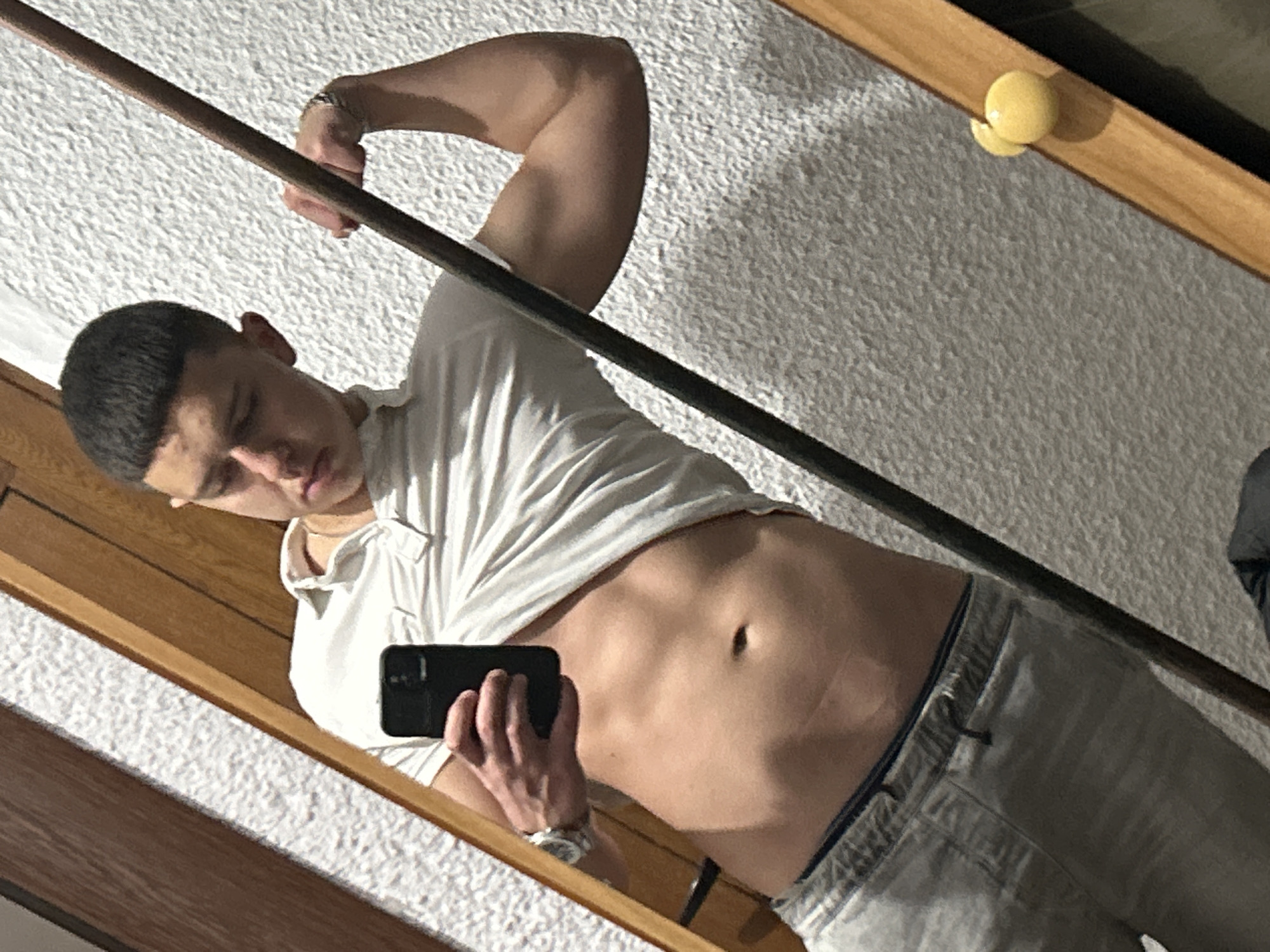 A selfie-style image captures a young man flexing his bicep while holding a smartphone. He is wearing a white t-shirt that is lifted to expose his abdomen and grey pants. A metal bracelet is visible on his left wrist, and a watch on his right. The background suggests an indoor setting with textured white walls. The image is a mirror reflection, angled within a wooden frame. There is no information to suggest this image was taken in the city of Zlatograd, Bulgaria.