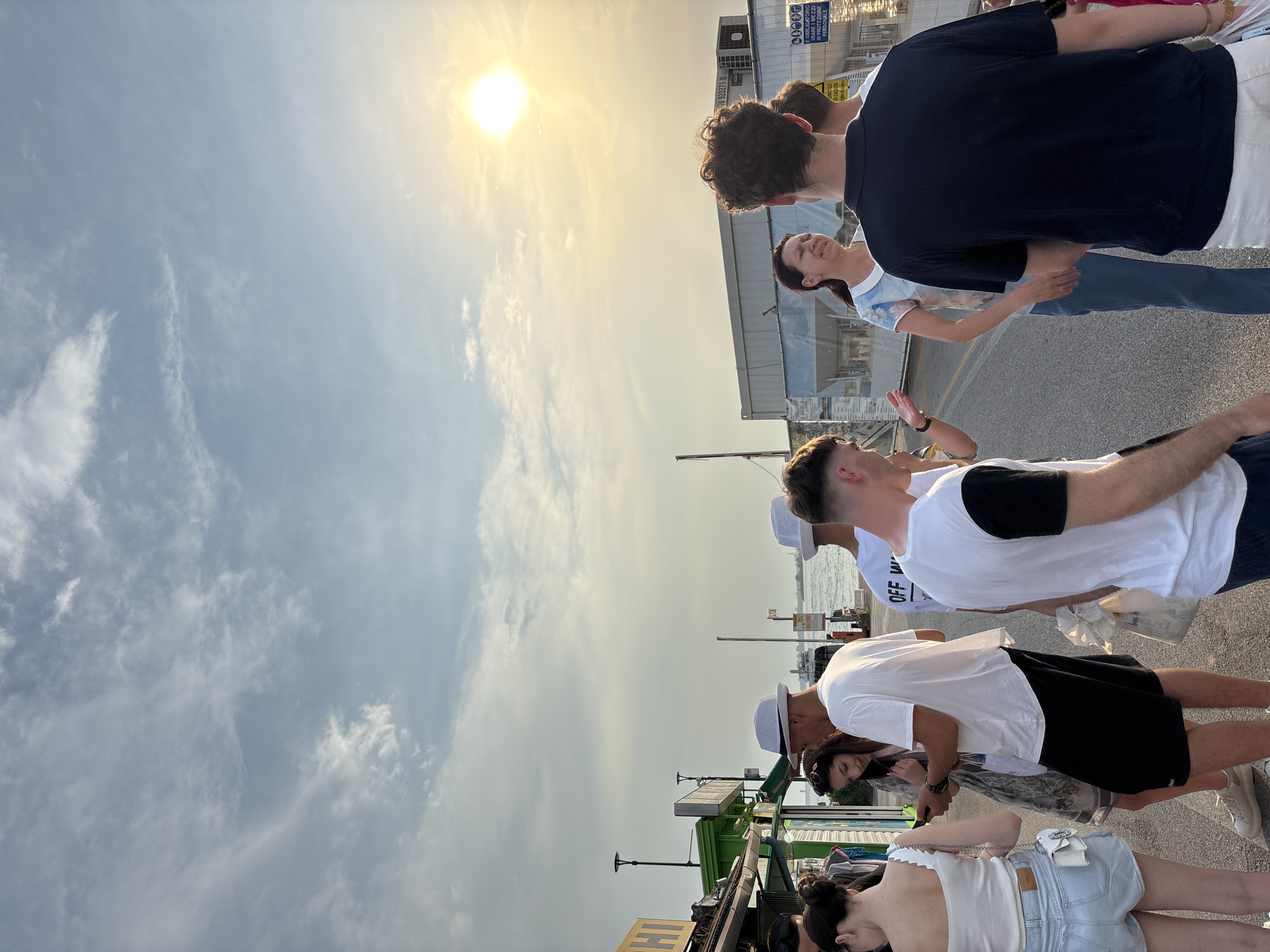 The image shows a group of people standing near what appears to be a bus stop in Punta Sabbioni, Italy. The location has a view towards the sea and sky. The people are dressed casually. The sun is visible in the sky, and there are some clouds. The perspective of the photo is taken from a slightly higher viewpoint, looking downward at the group.