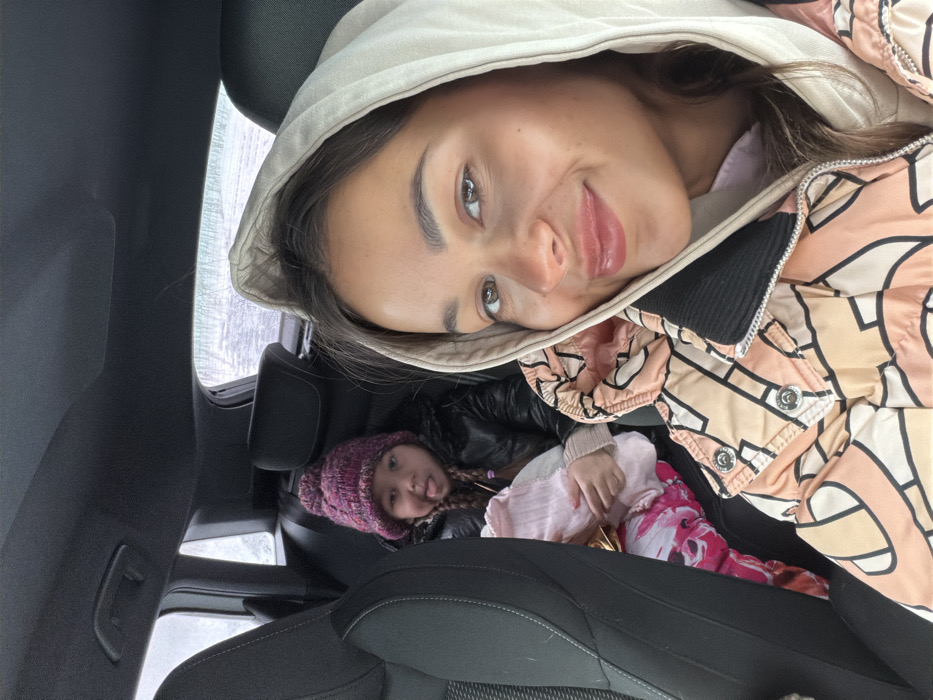 Inside a car, a woman wearing a hooded jacket poses for a selfie. In the back seat, a young girl in a purple knit hat is nestled under a black jacket. Outside the car window, bare trees and a snowy landscape are visible. The interior features black car seats and panels. The overall composition suggests a cozy, family road trip scene.
