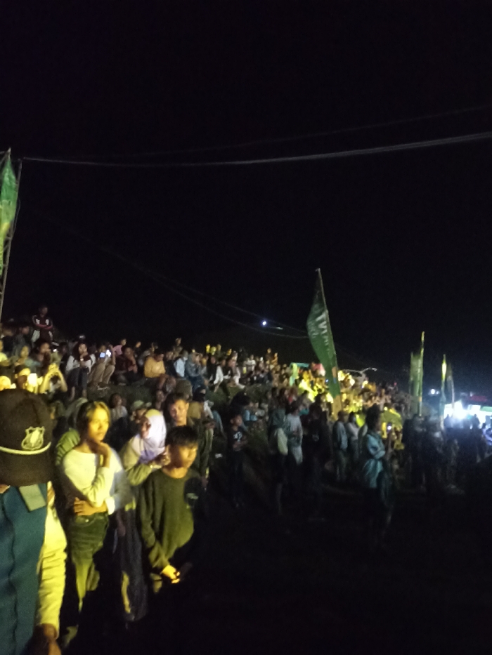 The image depicts a large outdoor gathering of people at night in Kecamatan Pujut, Indonesia. A dense crowd, composed primarily of young adults and some children, occupies an uneven landscape, possibly a hillside or natural incline, with many individuals standing or sitting.
The scene is illuminated by artificial lights, casting bright glows on sections of the crowd while leaving other areas in deep shadow, consistent with a nighttime event. The dark sky overhead shows subtle traces of power lines or cables. Several green flags are visible throughout the crowd, mounted on poles. One prominent flag clearly displays the text "TVRI," indicating an association with Televisi Republik Indonesia. Many attendees are looking in the same direction, suggesting a focal point such as a stage or screen is present but outside the frame. Some individuals are seen holding up their smartphones, possibly recording or photographing the event. In the left foreground, a person wearing a dark uniform, including a cap with a badge, is partially visible, potentially indicating security or event staff. The overall atmosphere suggests an engaged and attentive audience at a public event.