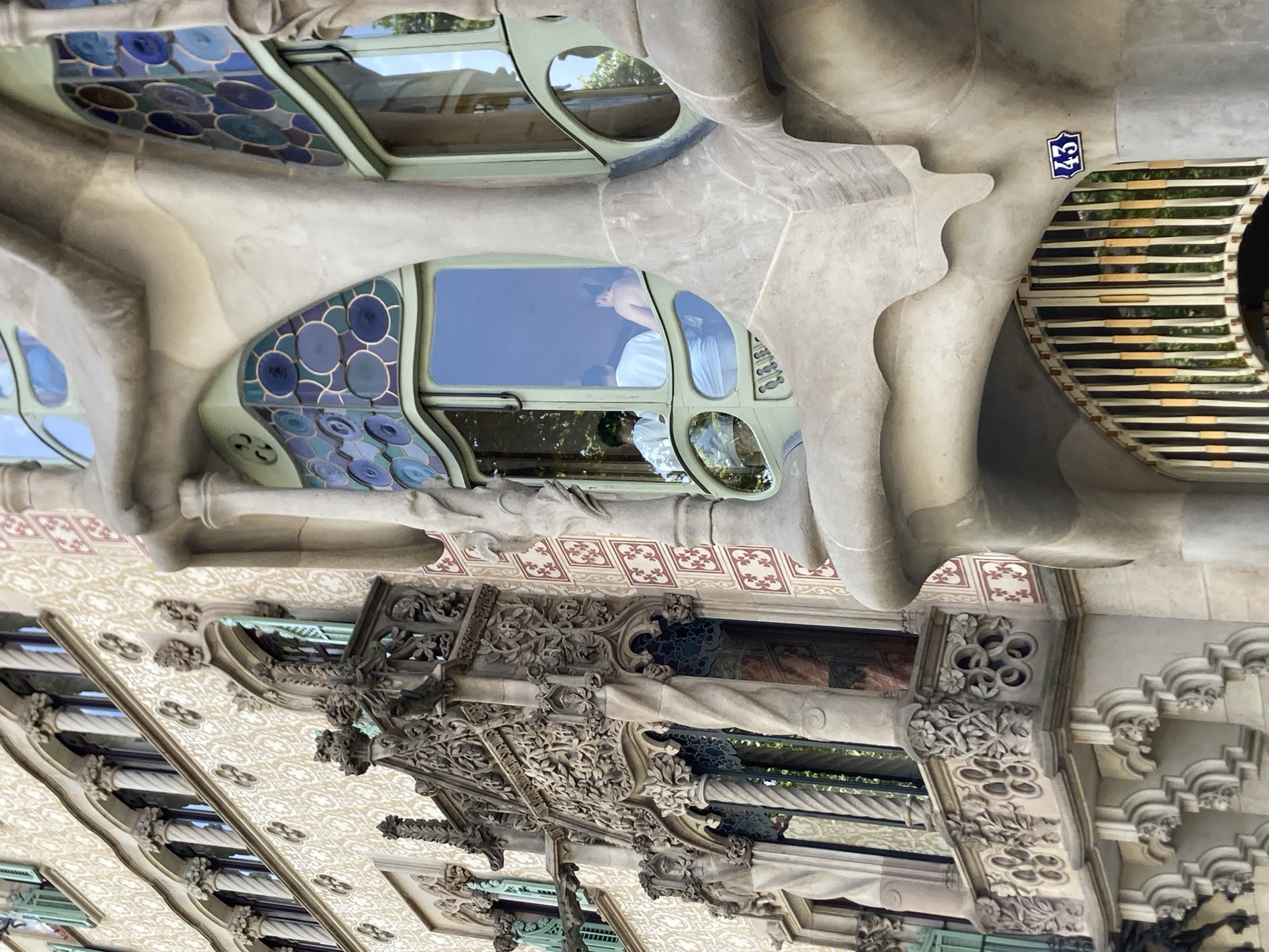 This image shows an external view of the Casa Batlló in Barcelona, Spain, with its distinctive organic architecture. The building's facade is characterized by undulating stone structures and colorful mosaic details, particularly around the windows. One window features an intricate design of circular blue and green stained glass. Another prominent window, framed in a light green material, offers a view into the interior where a person wearing a light blue shirt is visible, looking out. The building's ornamentation includes detailed stonework, reminiscent of floral and natural forms, and patterned tiles. A small blue plaque with the number "43" is affixed to the facade. The scene is bathed in natural light, suggesting it was taken during the daytime. The overall impression is one of artistic and architectural creativity, typical of Antoni Gaudí's work.
