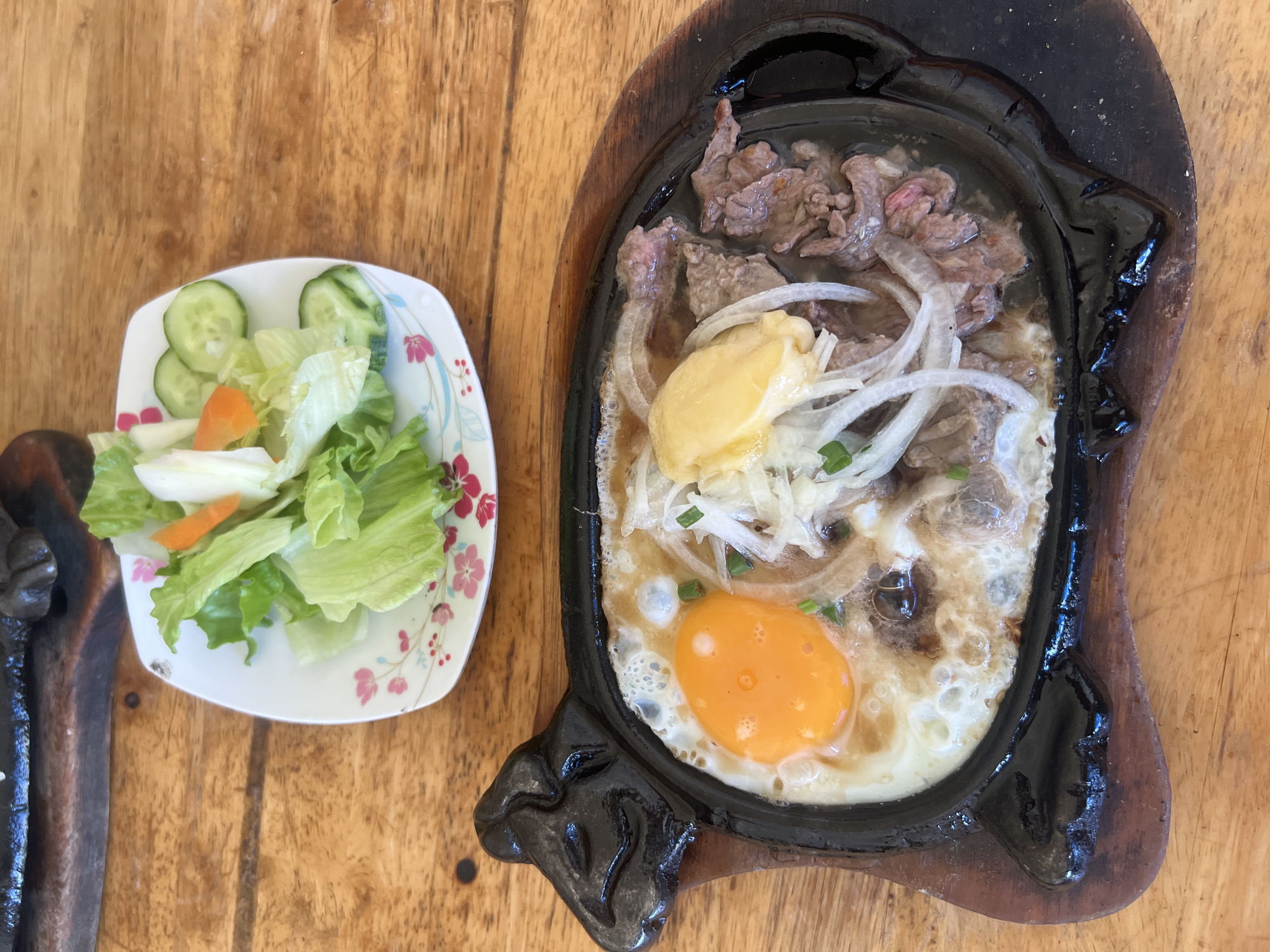 An overhead view presents a Bò Né (sizzling beef and egg) meal served on a rustic wooden table, indicative of an eatery in Nha Trang, Vietnam. The main dish occupies a black, cow-shaped cast-iron platter resting on a wooden base. It contains seared beef strips, a single sunny-side-up egg with a vibrant orange yolk, thinly sliced white onions, and a small pat of yellow butter, all actively sizzling. A separate small, square white plate with a pink floral motif holds a side salad composed of fresh green lettuce leaves, cucumber slices, and a small piece of orange carrot. The light brown wooden table surface shows natural grain and appears worn.