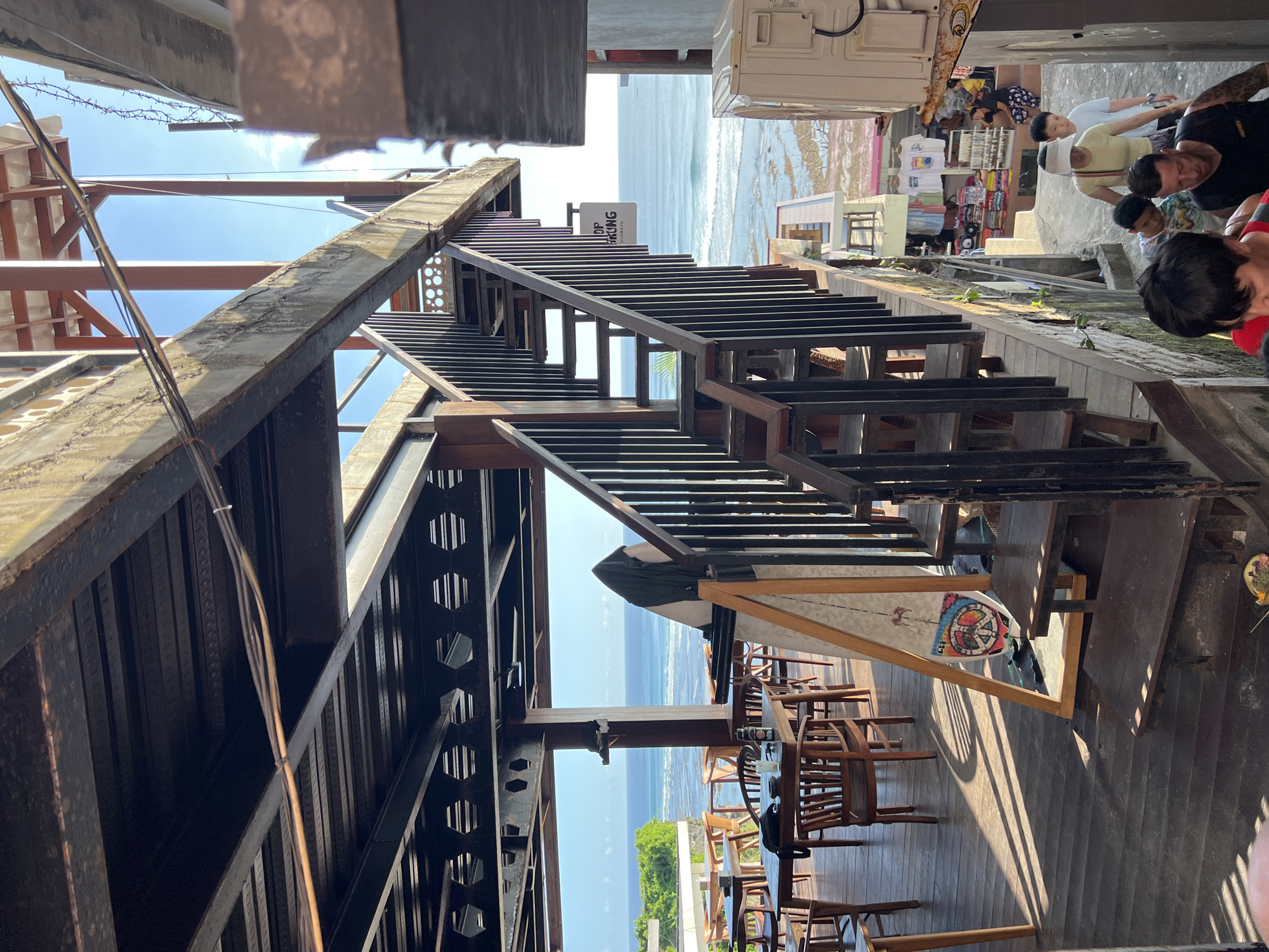 Here is a detailed description of the image:
The image shows an outdoor scene, likely a restaurant or lounge area, with a view of the ocean. The location appears to be multi-tiered, with wooden stairs leading down towards the water. The stairs are constructed with dark metal supports and wooden steps.
On the lower level, there is an arrangement of wooden tables and chairs, suggesting a dining area. A surfboard stands propped against a railing. Overhead, a structural support system with metal beams and a partial roof is visible.
In the background, the ocean stretches to the horizon under a clear sky. Along the shoreline, there is evidence of a beach area with people and small buildings. A few individuals are visible in the top right, appearing to be tourists or visitors in a shopping area.