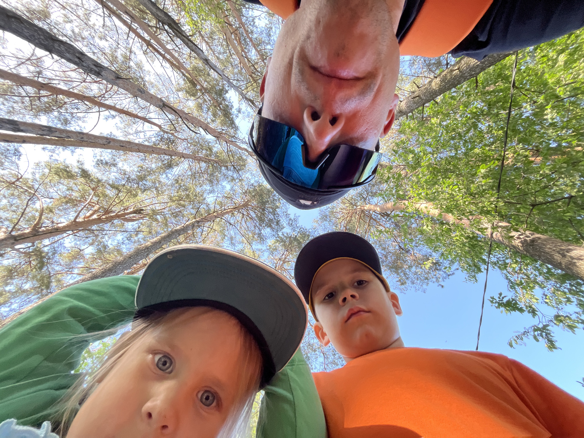The image depicts a low-angle selfie taken from below, showing a man and two children. The man, at the top, wears sunglasses and a cap, and is looking down at the camera. To the right, a child in an orange shirt and a black cap is also looking down. To the left, another child wearing a green jacket and a cap is gazing upwards. The background consists of tree branches and leaves, suggesting a forested area. The presence of coniferous trees points to a location like a park or forest in Chaikovsky, Russia, where such landscapes are common. The picture seems to be an informal shot capturing a moment of family or group activity outdoors.