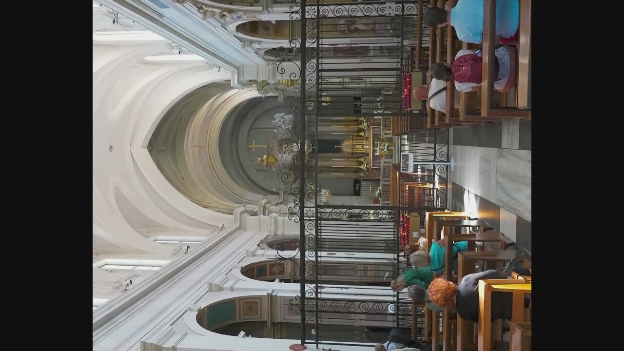 The image depicts the interior of a church in Morlupo, Italy. The central nave is visible, with rows of wooden pews on either side. A wrought-iron gate separates the congregants from the altar area. The altar itself is elaborate, featuring a large golden structure with a cross at its apex, flanked by statues and candles. The ceiling is vaulted and painted white, with arched windows running along the upper walls. Several people are seated in the pews, facing the altar. Some are looking forward, while others are engaged in conversation. The overall atmosphere is one of solemnity and reverence.