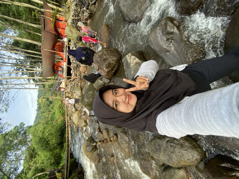 A young woman wearing a black hijab, a grey long-sleeve shirt, and black pants sits on rocks near a stream, flashing a peace sign at the camera. Behind her, a flowing stream cuts through a green, lush landscape. Some people are visible in the background. A small wooden bridge crosses the stream further back. Tents are visible in the background.