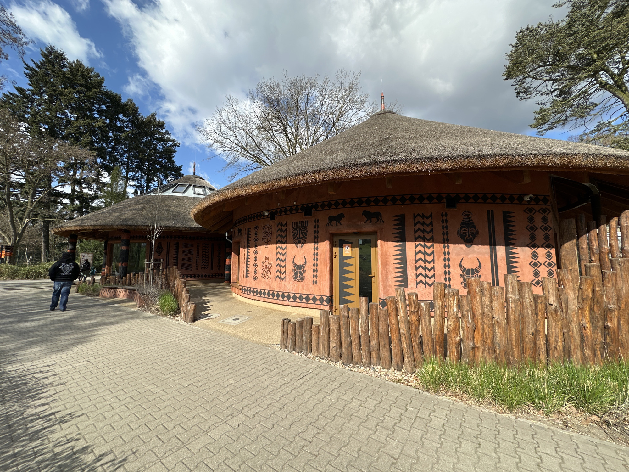 The image depicts a pathway leading to two circular buildings with thatched roofs, likely part of a zoo exhibit. The buildings are decorated with traditional African-inspired artwork, featuring geometric patterns, depictions of animals, and human faces. A low wooden fence lines part of the pathway adjacent to the buildings. A person in casual clothing is walking along the pathway toward the first building. The scene is set on a sunny day with a partly cloudy sky. Based on the architectural style and available geographical information, the location is likely the Wrocław Zoo in Wrocław, Poland, which features an African habitat exhibit.